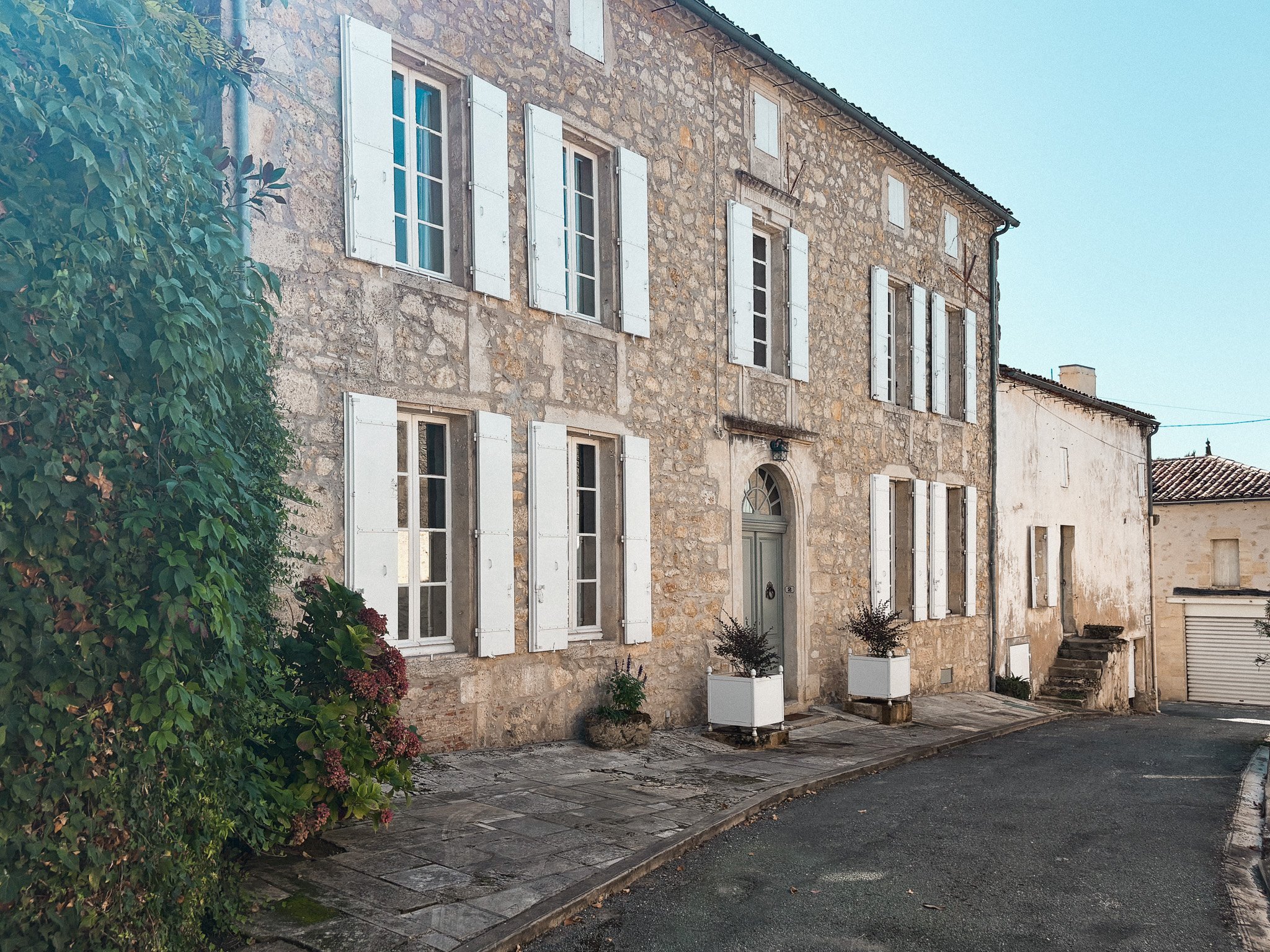 A stone building with white shutters and multiple windows, two potted plants at the entrance, and a winding paved sidewalk in front, with a neighboring building and a garage door to the right.