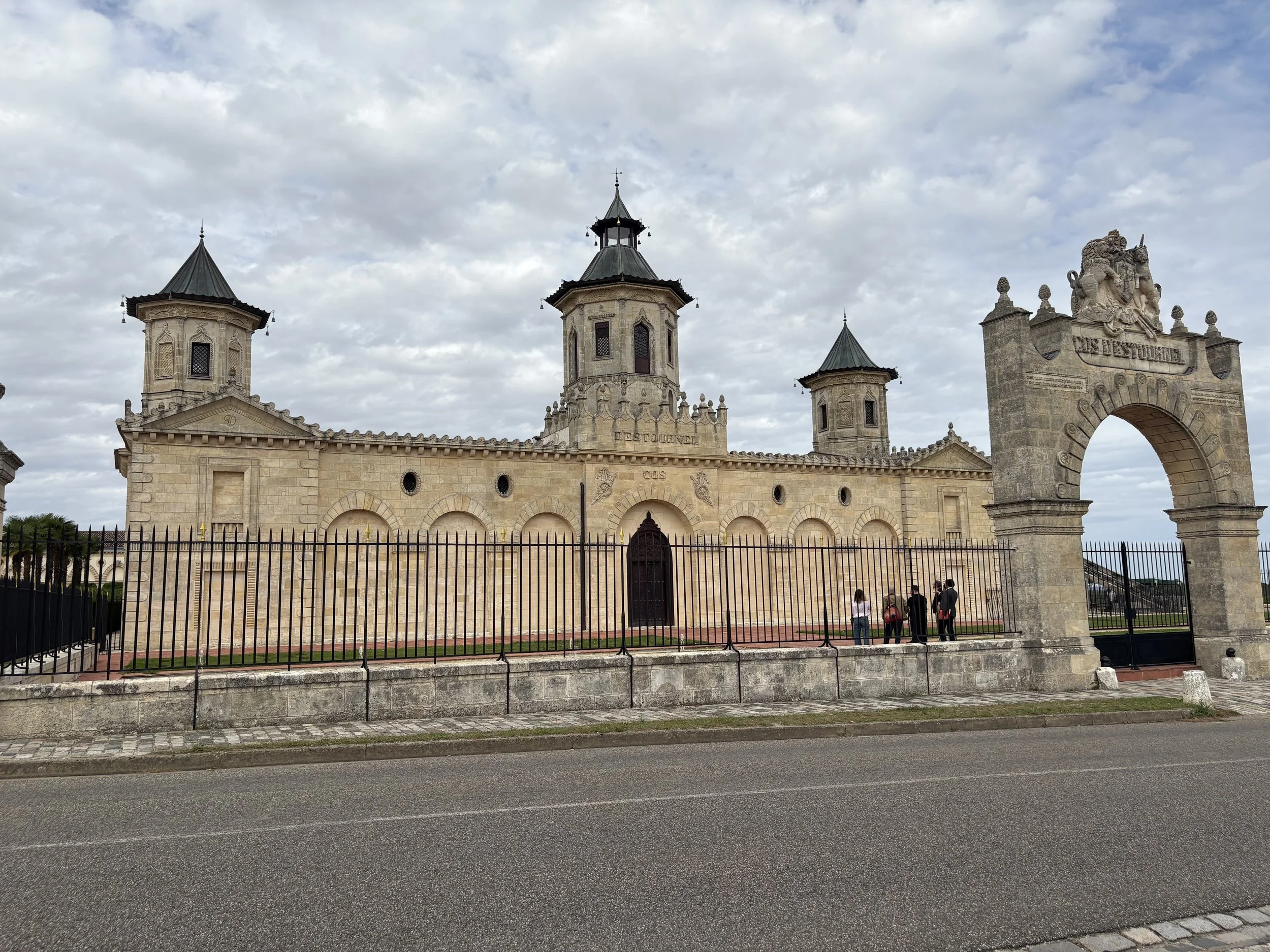 Historical stone fortress with three towers and an ornate arched entrance, surrounded by a metal fence, with people walking near the entrance and a cloudy sky overhead.