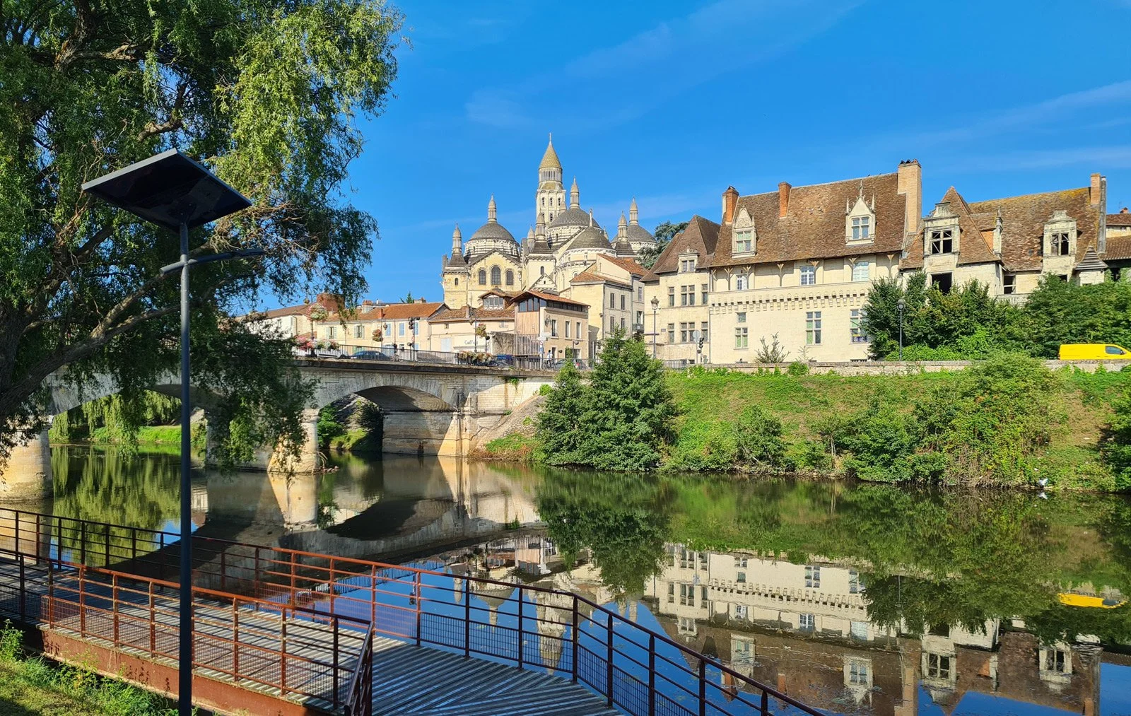 A scenic view of a European town with historic buildings and a castle on a hill, reflected in a calm river beneath a stone bridge on a clear blue day.