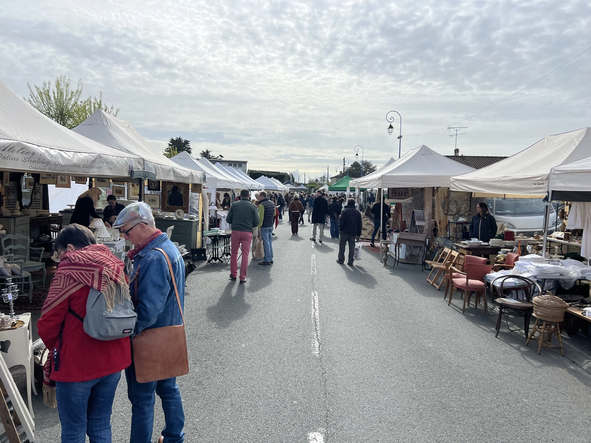 Outdoor street market with white tents and vendors selling various items, people browsing and walking on a cloudy day.