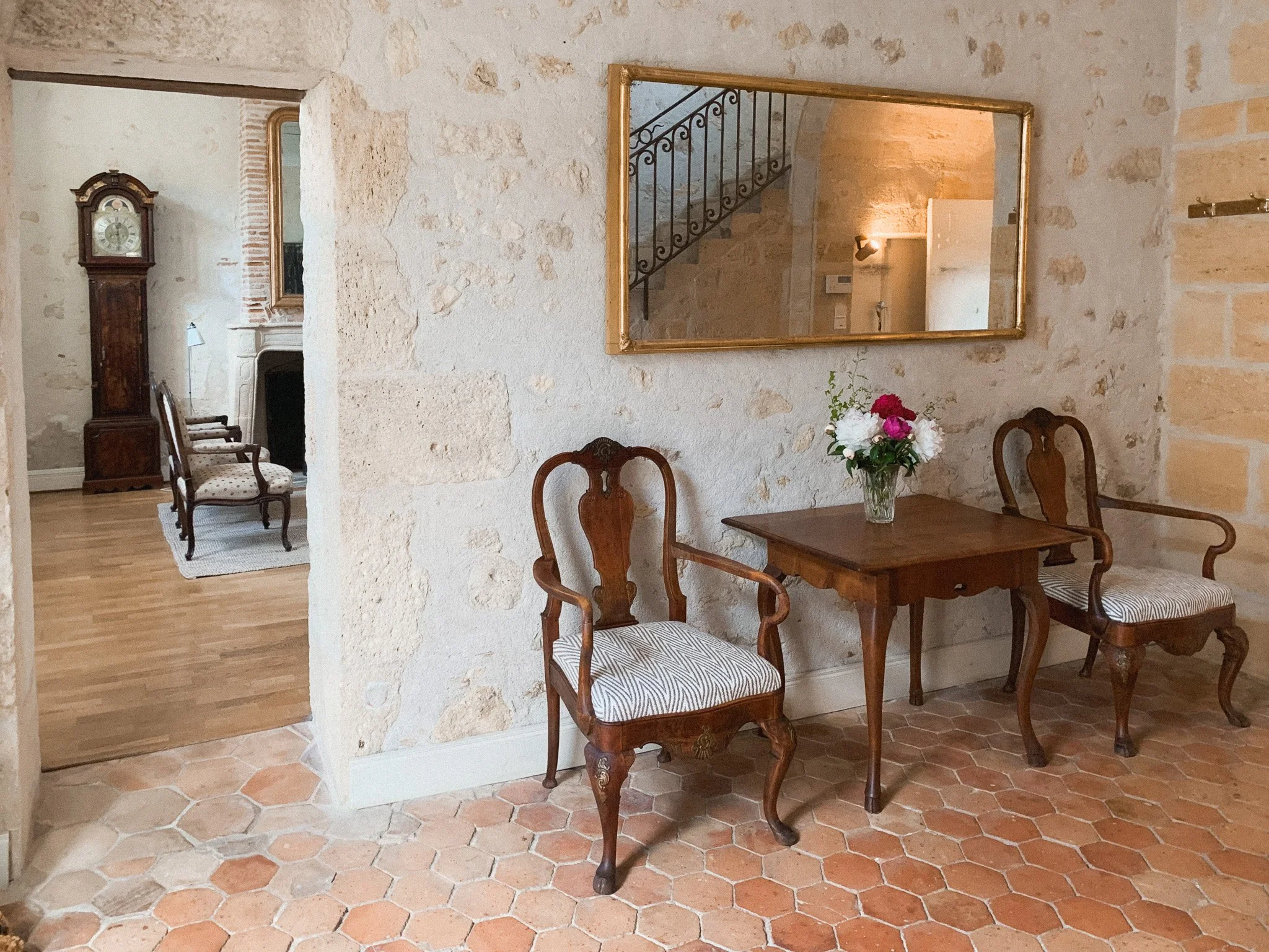 A cozy corner in a rustic home with two antique wooden chairs with padded seats, a small wooden table with a vase of pink and white flowers, a large rectangular mirror on the stone wall, and a view into another room with a grandfather clock and a fireplace.