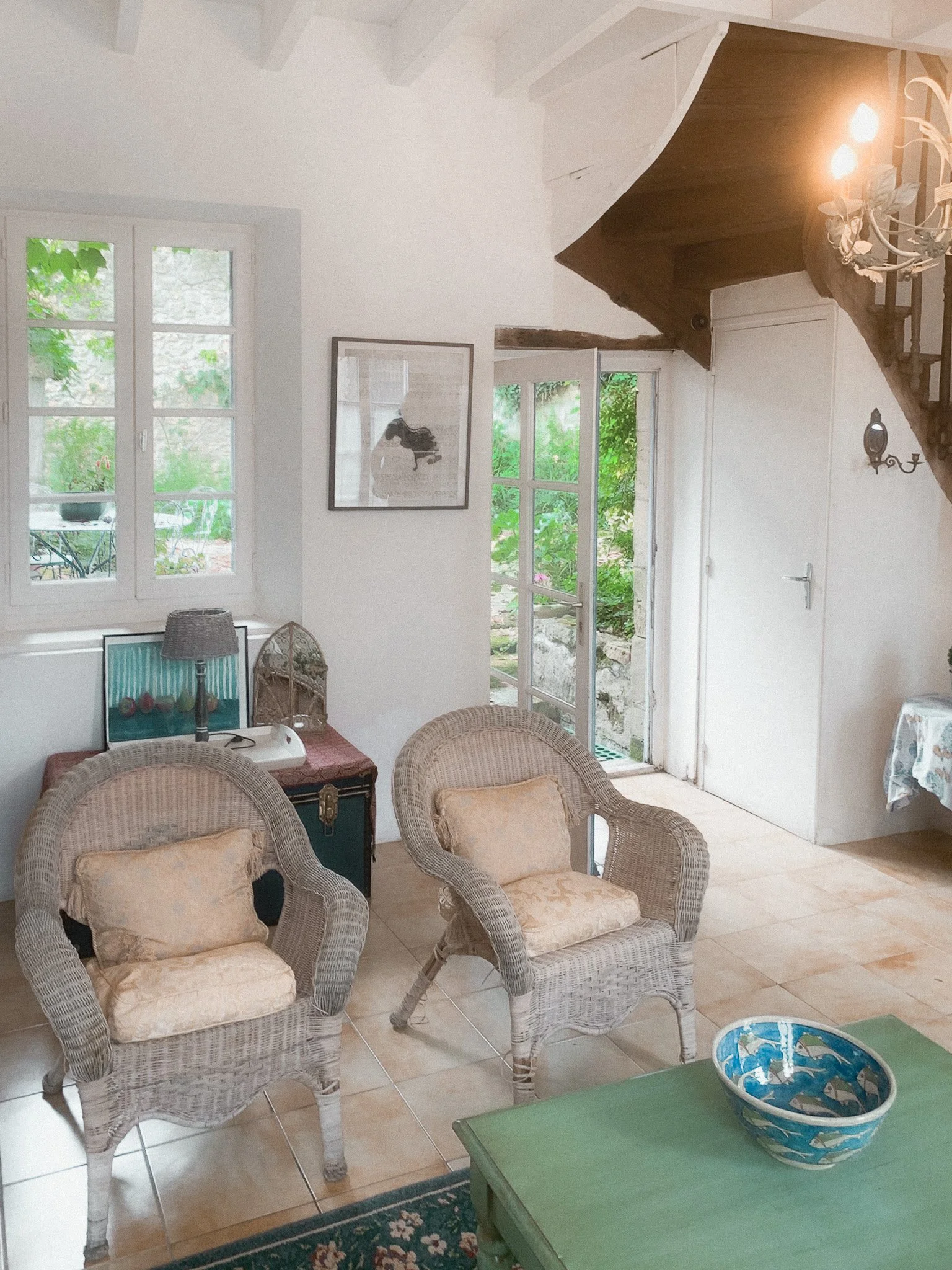 A cozy living room with two wicker chairs with beige cushions, a small green table with a blue bowl, and a large window and glass door leading to a garden outside. There is a framed picture on the wall, a small side table with a lamp, and a staircase