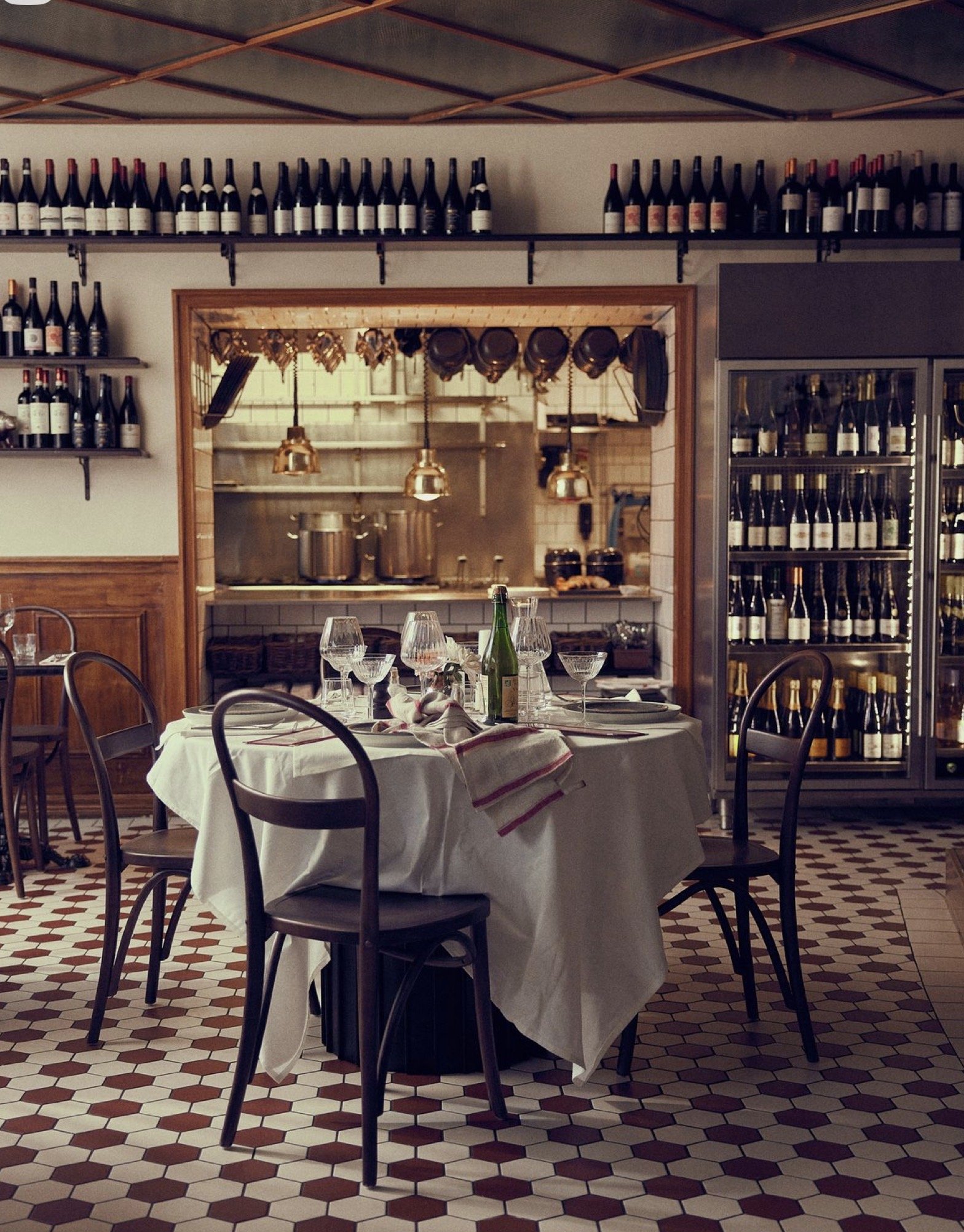 Restaurant table set with wine glasses, plates, napkins, and a bottle, with a wine storage area and kitchen visible in the background.