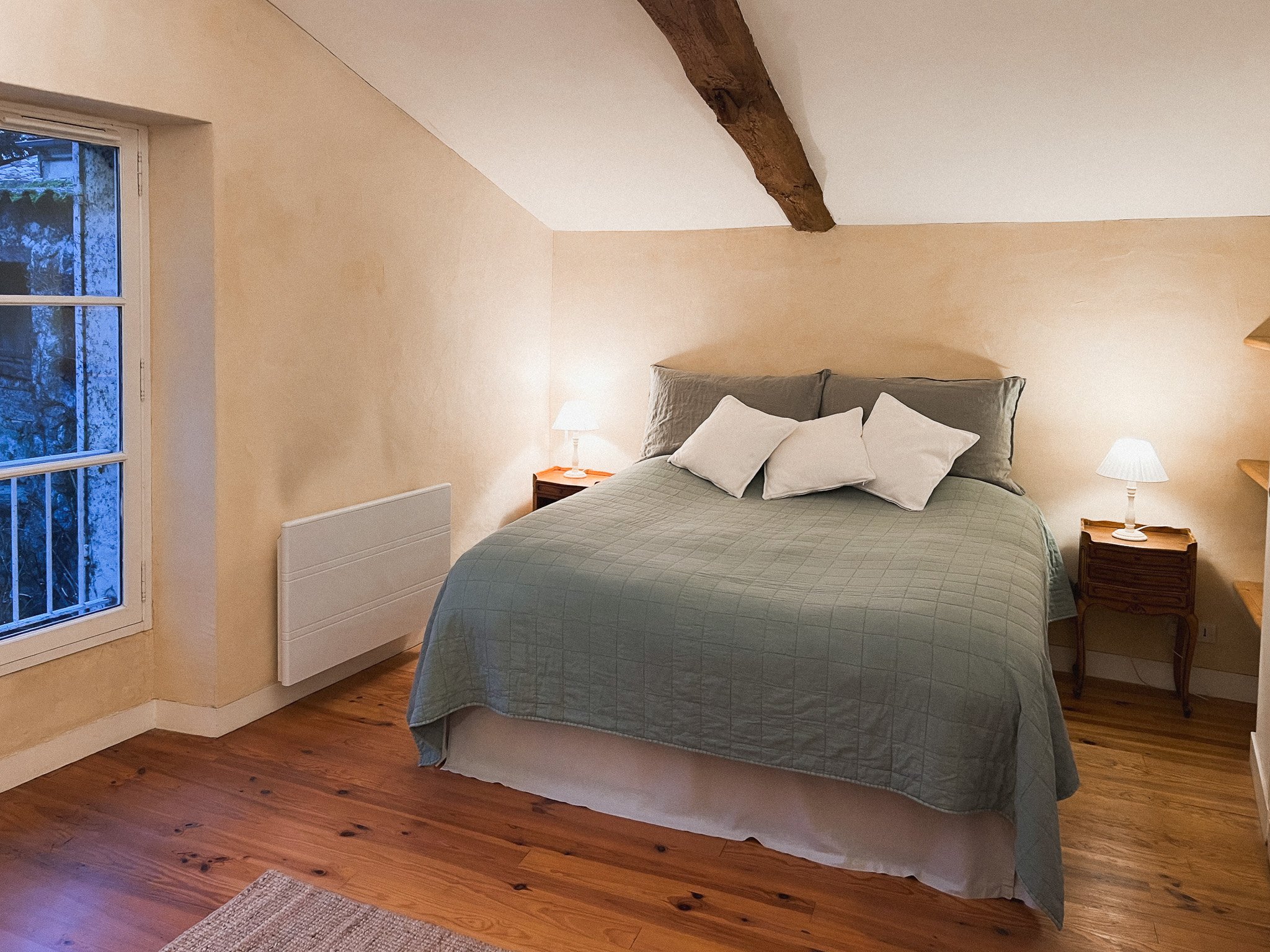 A cozy bedroom with a sloped ceiling, featuring a bed with a green quilt, white and gray pillows, bedside tables with lamps, a wooden floor, a window with a view of trees, and exposed wooden beams.