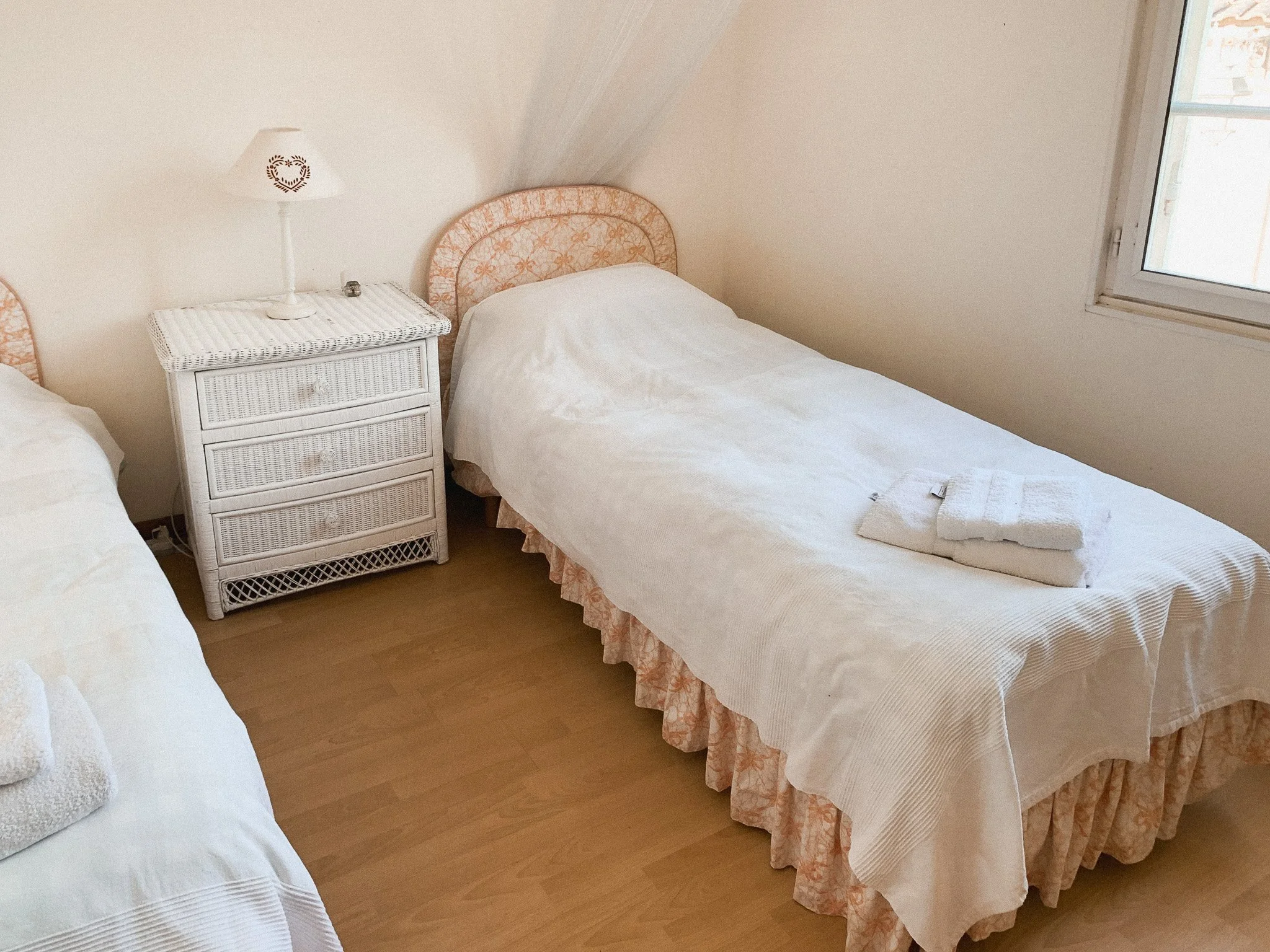 A bedroom with two single beds, a white wicker nightstand with three drawers, a lamp with a heart-shaped cutout, and folded towels on one bed. There is a window on the right side of the room.