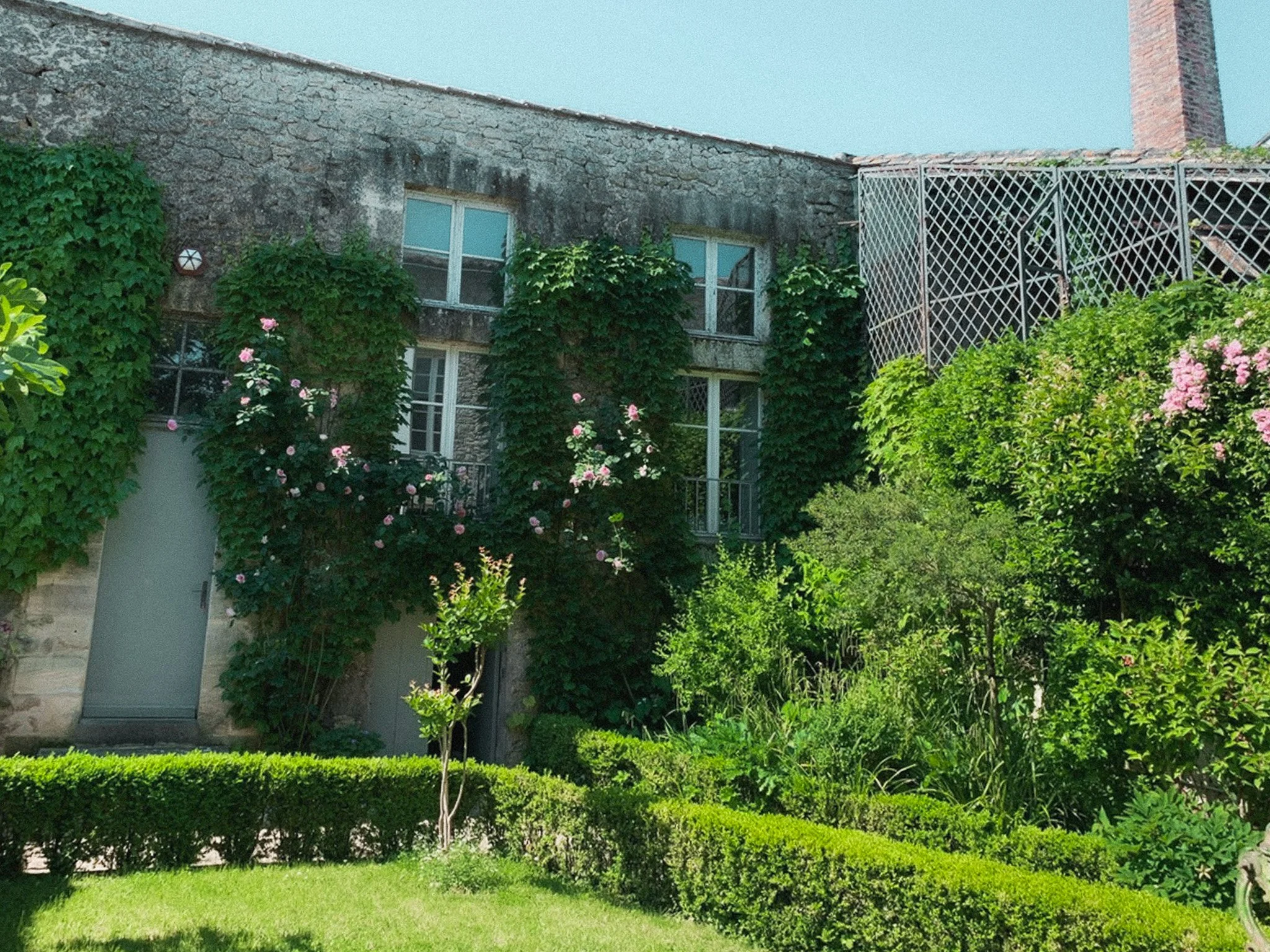 A stone house with ivy climbing up its walls, surrounded by a lush green garden with shrubs, small trees, and pink flowers, under a bright blue sky.