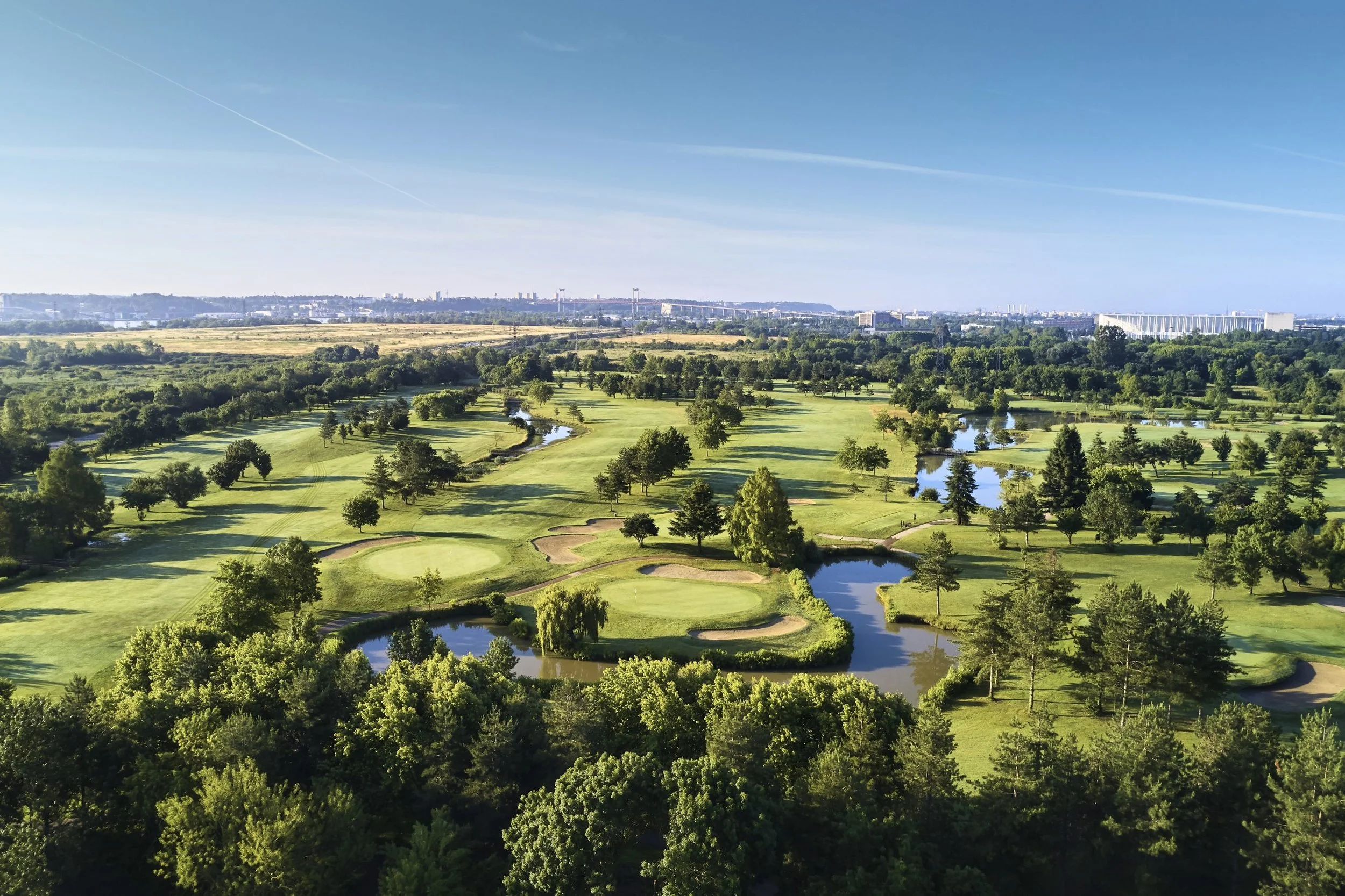 Aerial view of a lush green golf course with multiple water hazards, surrounded by trees, with city buildings and a bridge in the distance under a clear blue sky.