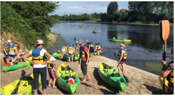 Group of people, including children, preparing to kayak on a river, with some on the shore and others paddling in the water.
