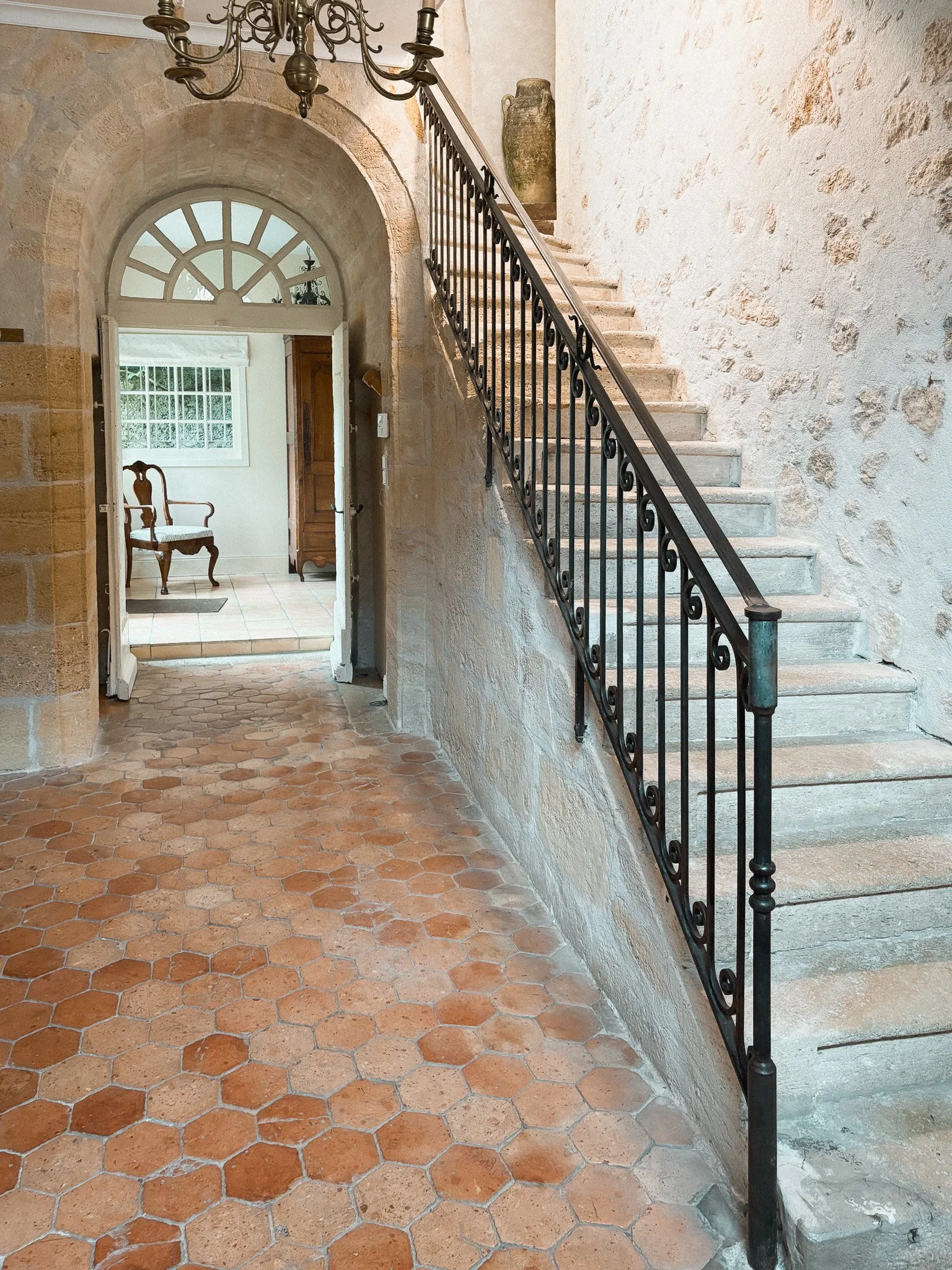 Interior view of a house with a tiled hexagon floor, stone walls, a staircase with a decorative wrought iron railing, and a chandelier hanging from the ceiling. There is an arched doorway leading to a room with a window and a chair.