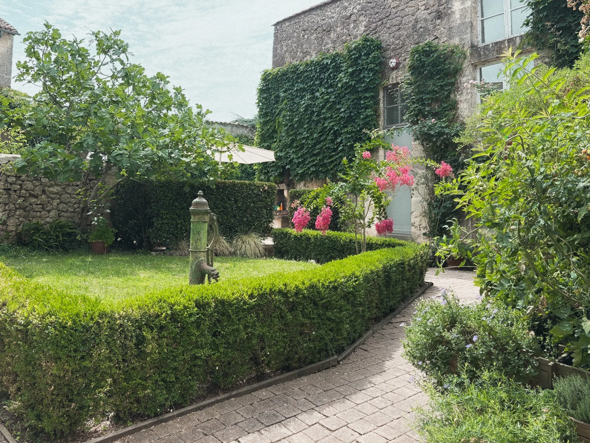 A lush garden with trimmed hedges, flowering plants, a stone pathway, and a stone building with ivy climbing its walls.
