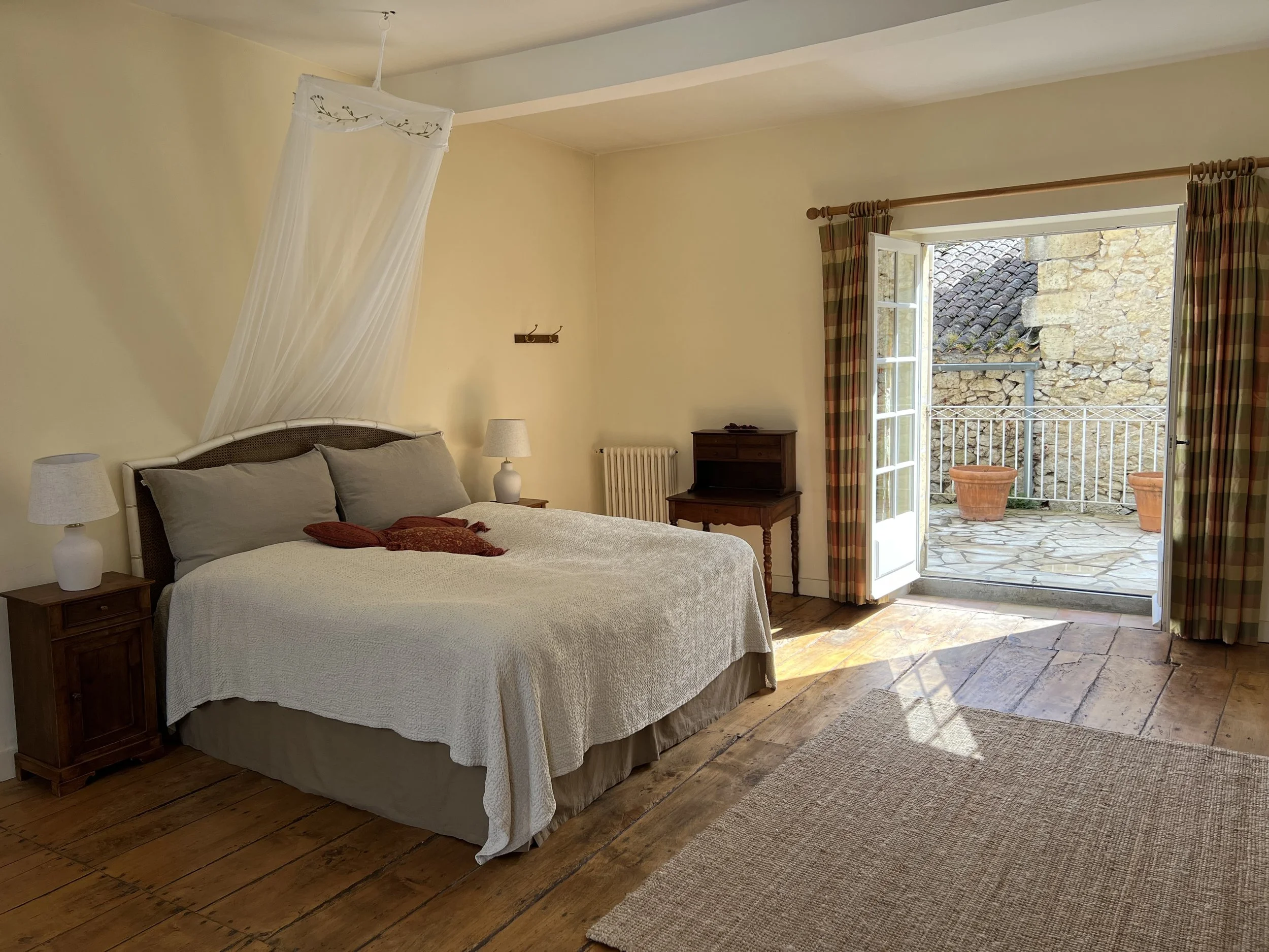 A cozy bedroom with a large bed, two nightstands with lamps, a small desk, and an open door leading to a stone-tiled balcony with two large planters. Sunlight streams in through the open door.