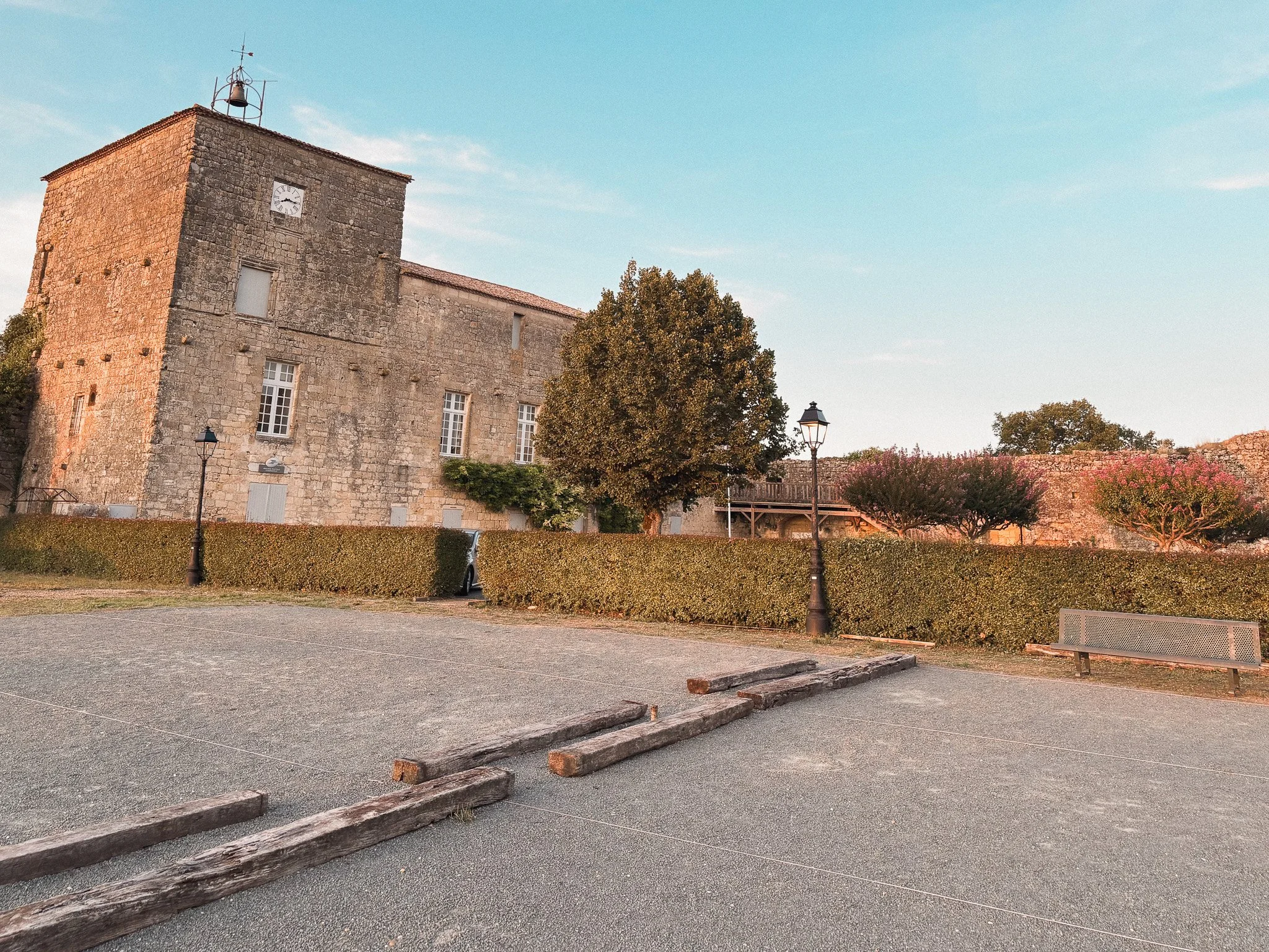 An old stone building with a clock tower, surrounded by trees, lamps, and a bench, with a gravel area and wooden posts in the foreground.