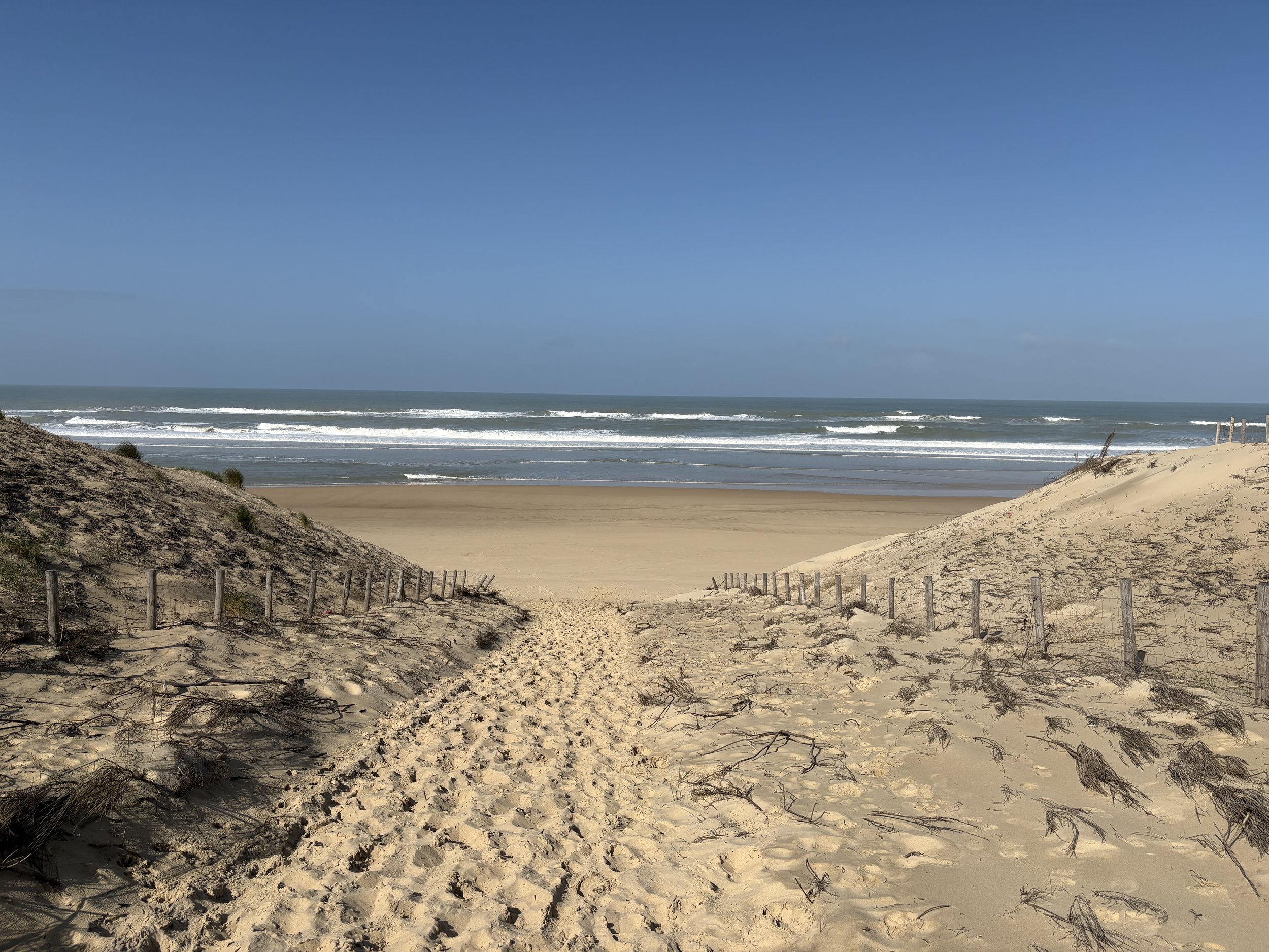 Sandy beach with tracks leading to the ocean, sand dunes on either side, and the sea with small waves under a partly cloudy blue sky.