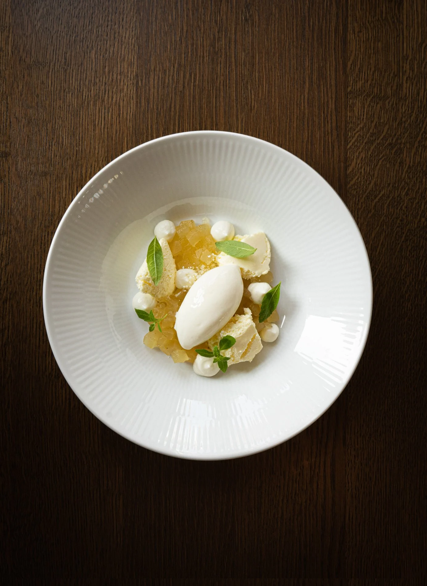 Dessert with white ice cream, yellow fruit compote, small white meringue dots, and green herbs on a white plate.
