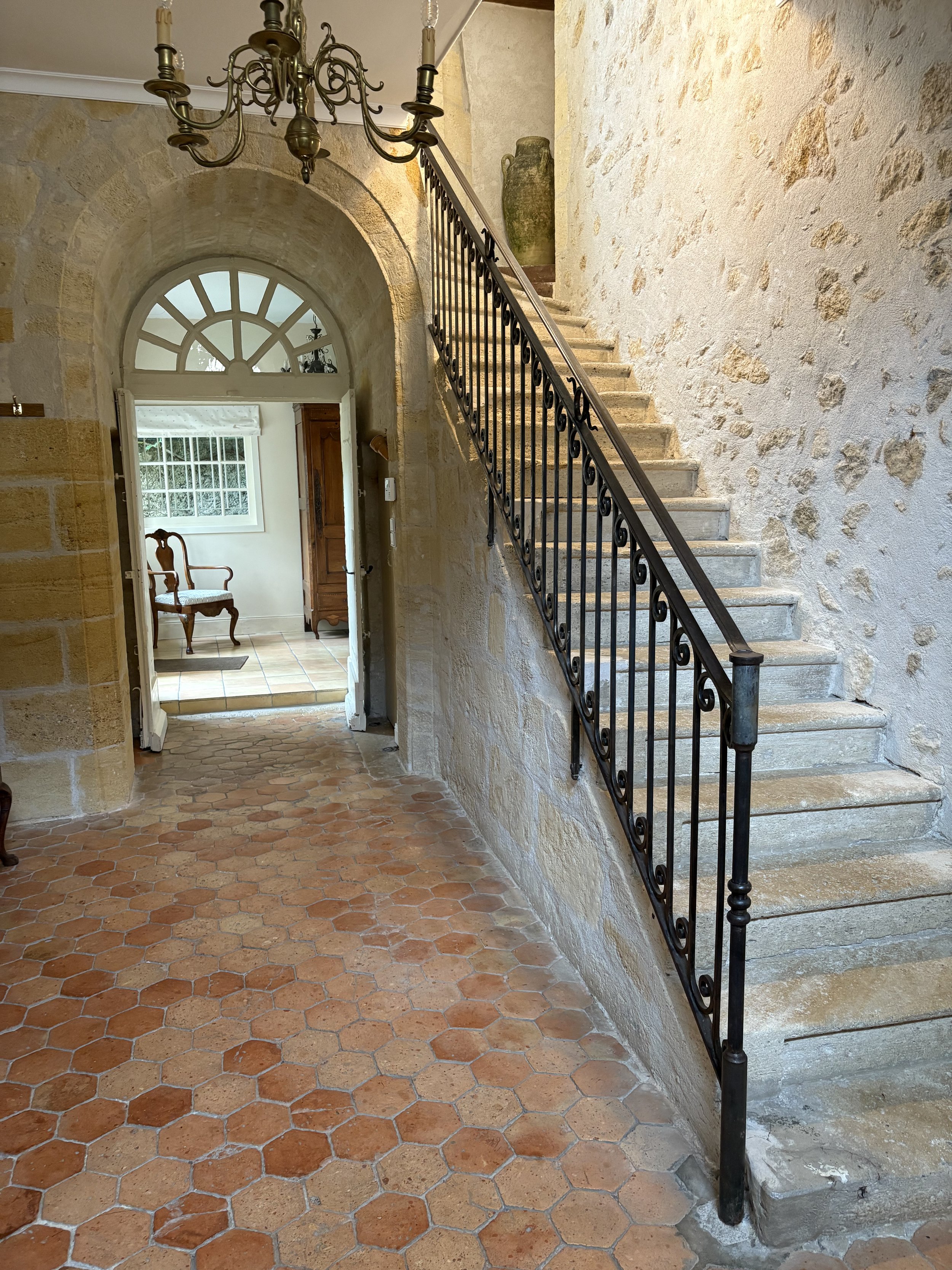 Interior of a rustic house with a stone staircase, an iron railing, terracotta tile flooring, a chandelier hanging from the ceiling, and a doorway leading to a room with a wooden chair, a large window, and a wooden cabinet.