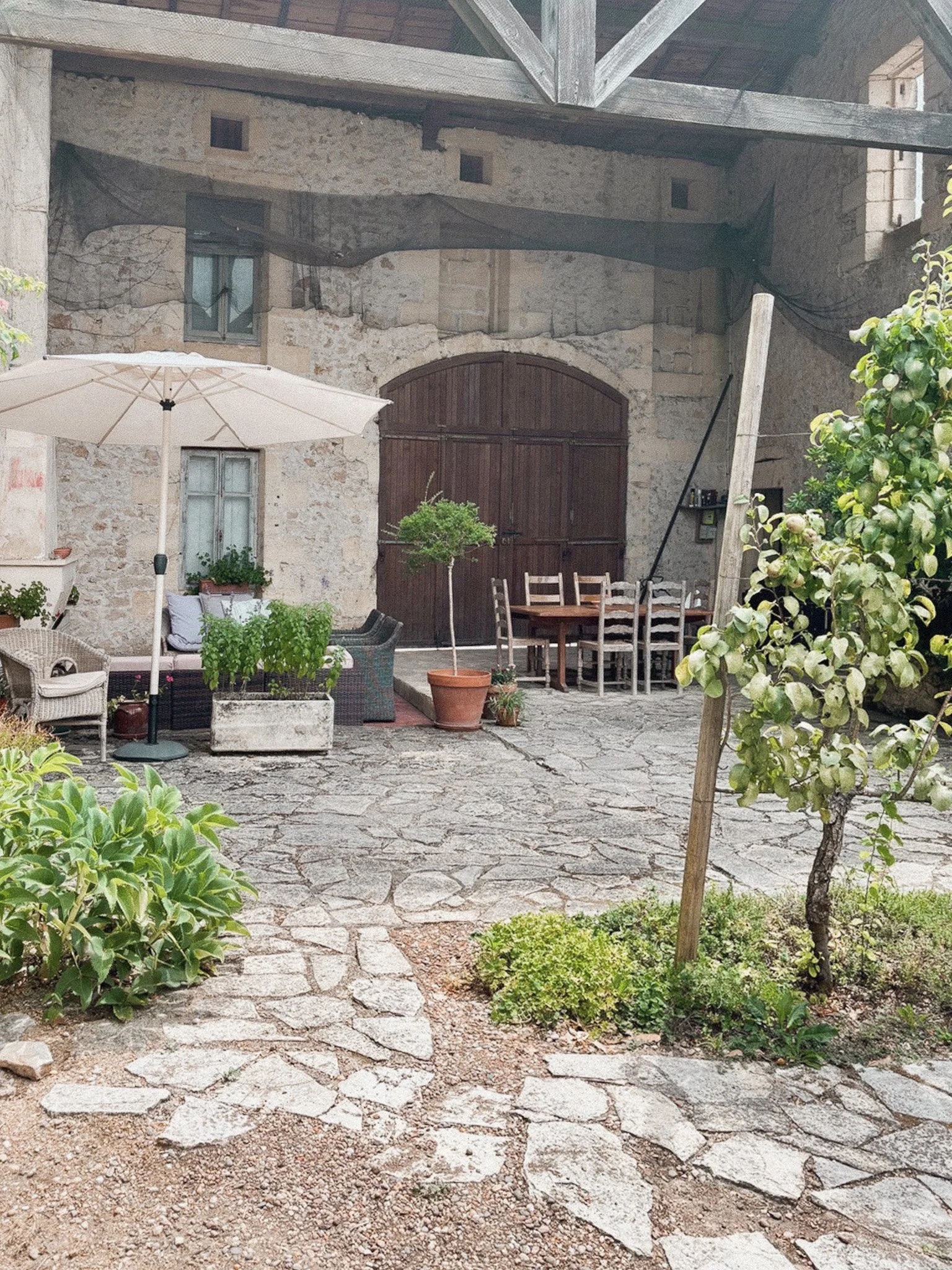 A rustic outdoor patio with stone pavement, green plants, a white patio umbrella, and outdoor furniture including a wicker chair and a wooden dining table with chairs. An old stone wall with wooden barns doors and a window is in the background.