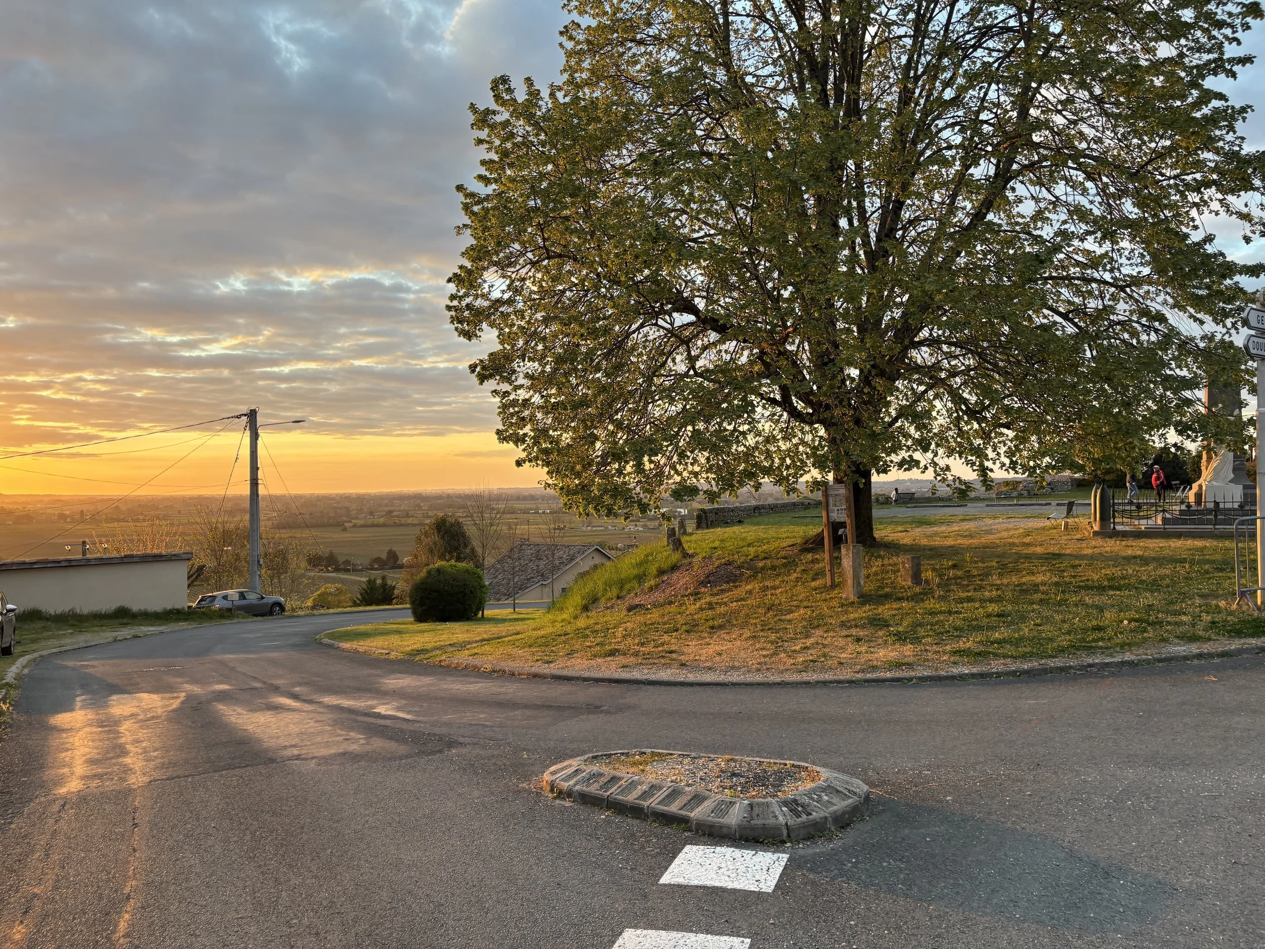 A scenic view of a small roundabout with a large tree in the center during sunset. The sky is partly cloudy with warm golden hues, and in the background are distant fields and mountains. There are a few cars and some pedestrians near a monument or st