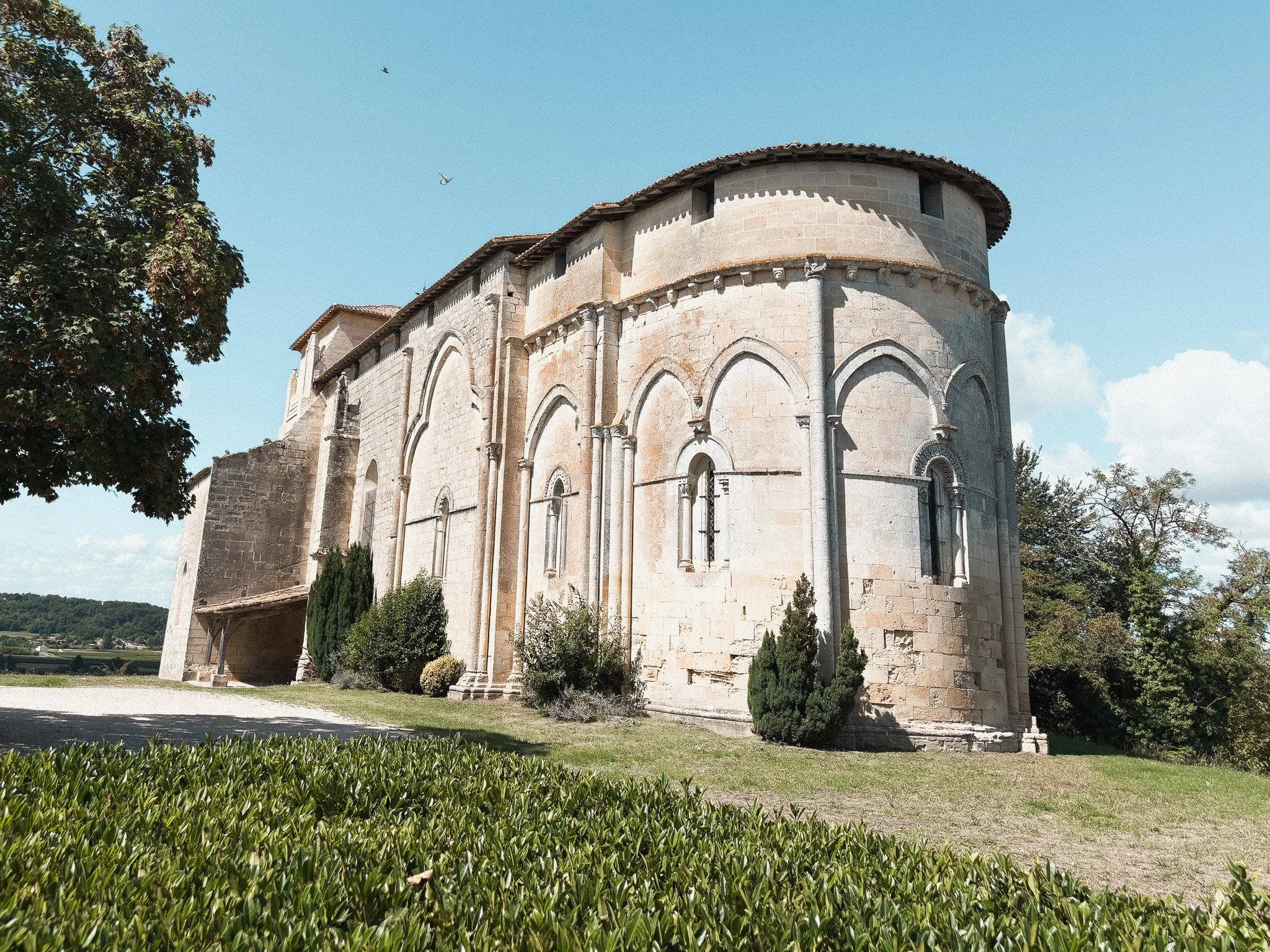 A historic stone church with arched windows and a rounded tower, surrounded by greenery under a blue sky with scattered clouds.
