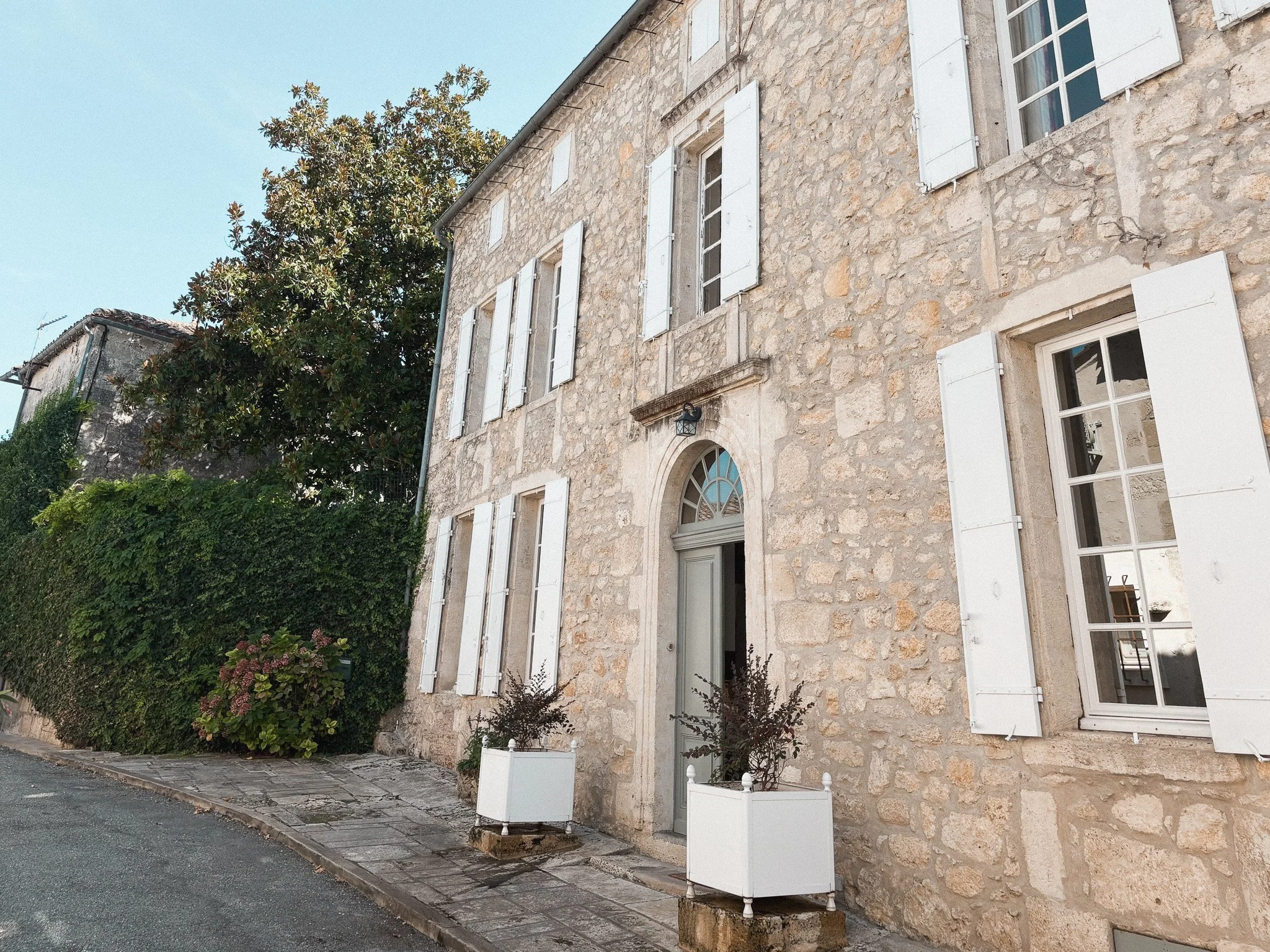 A stone house with white shutters on windows, a grey door, and potted plants outside. There are bushes and a tree beside the house, and the sky is clear