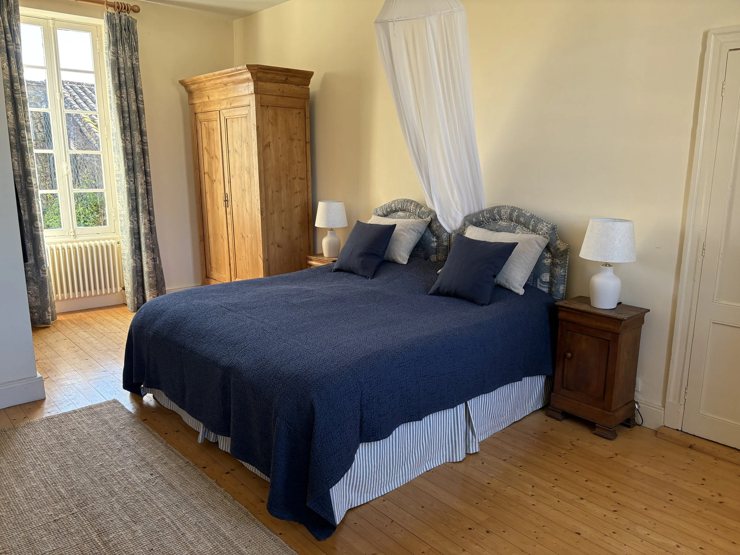 A bedroom with a wooden wardrobe, a bed with a blue bedspread, four pillows, two nightstands with white lamps, a window with curtains, and hardwood floors.