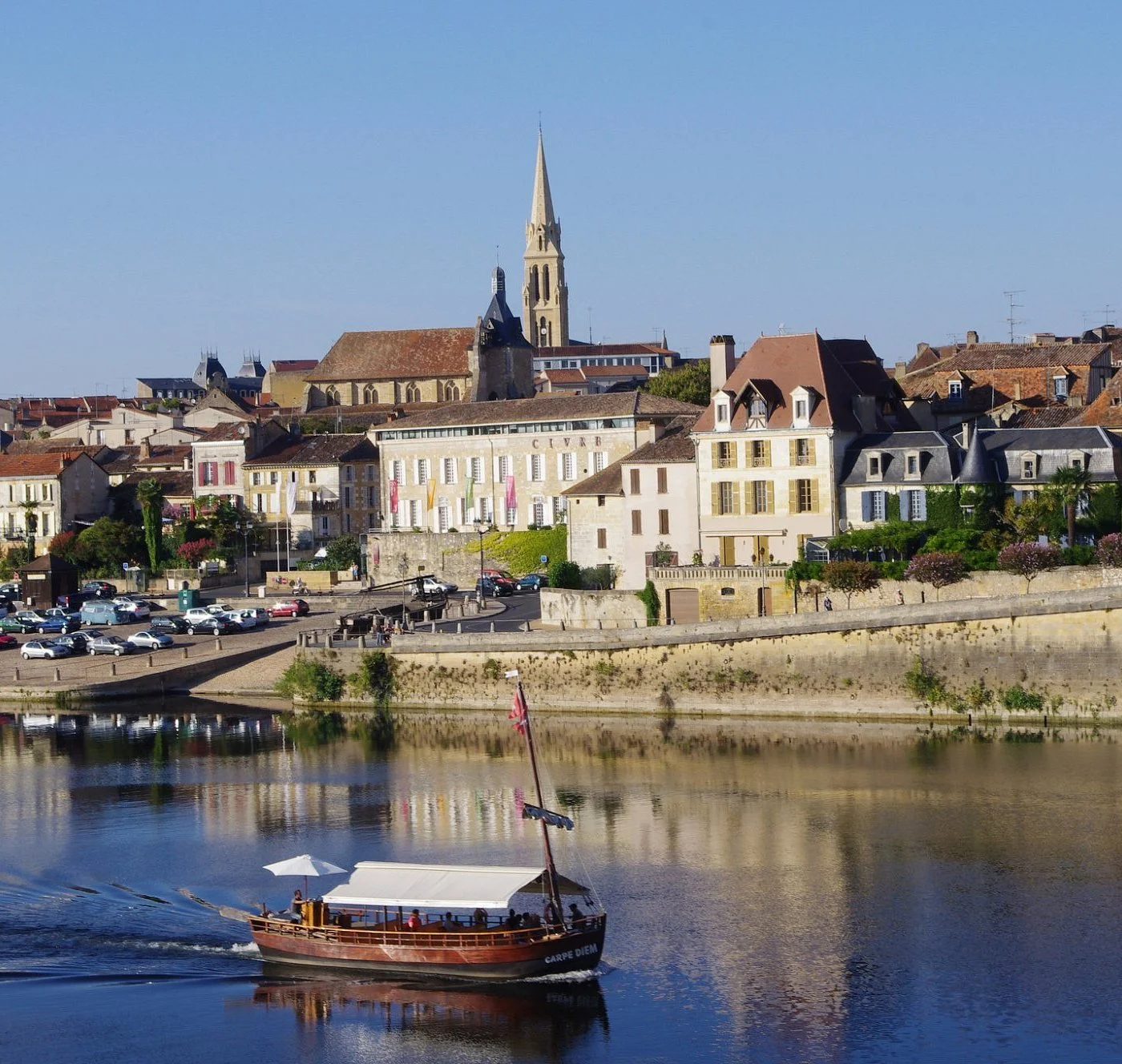 A river with a wooden boat and a white canopy sailing past a European town with historic buildings and a church with a tall steeple, set against a clear blue sky.