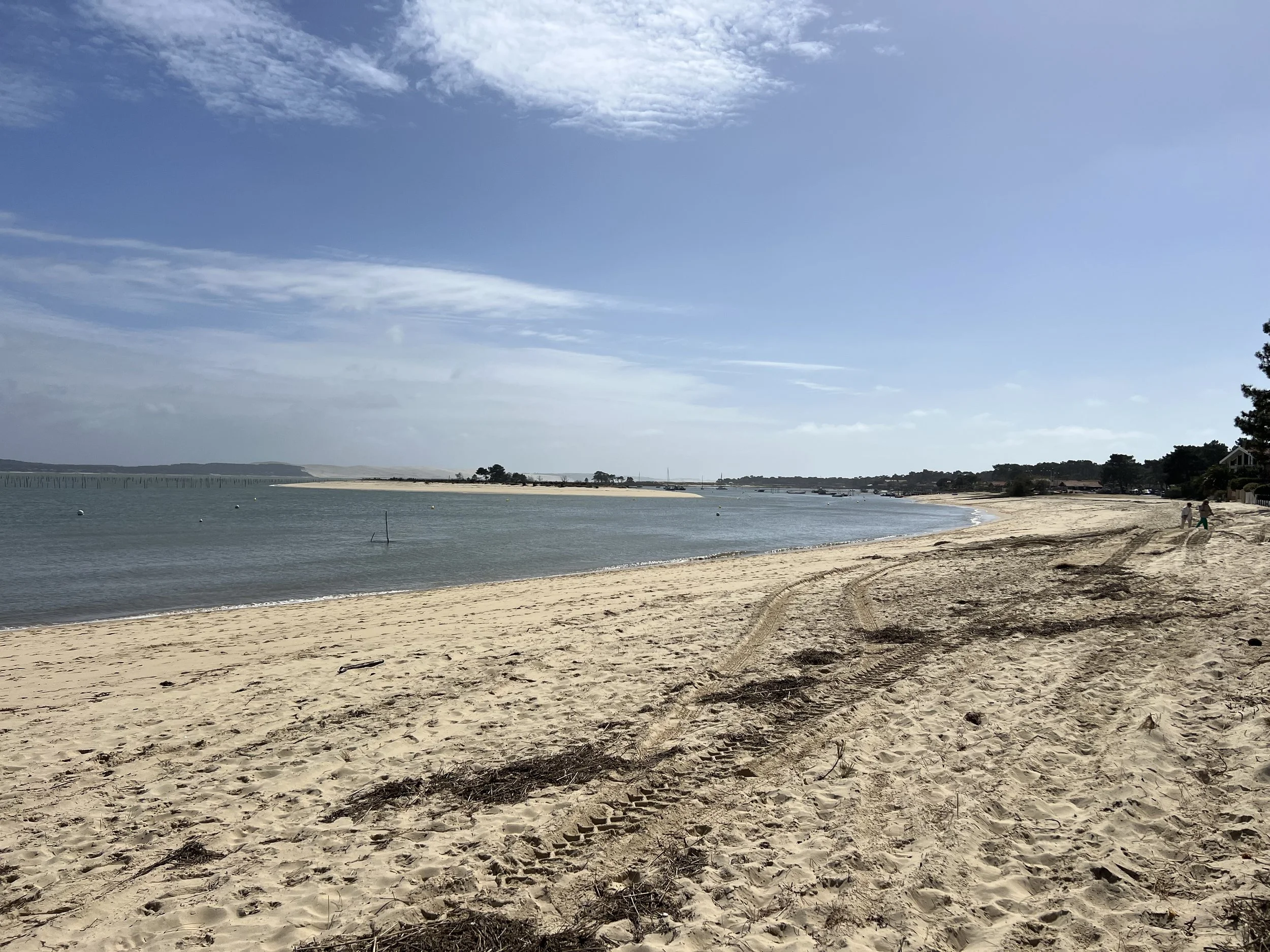 A sandy beach next to calm water with tire tracks in the sand, a few people walking, and houses along the shoreline under a partly cloudy sky.