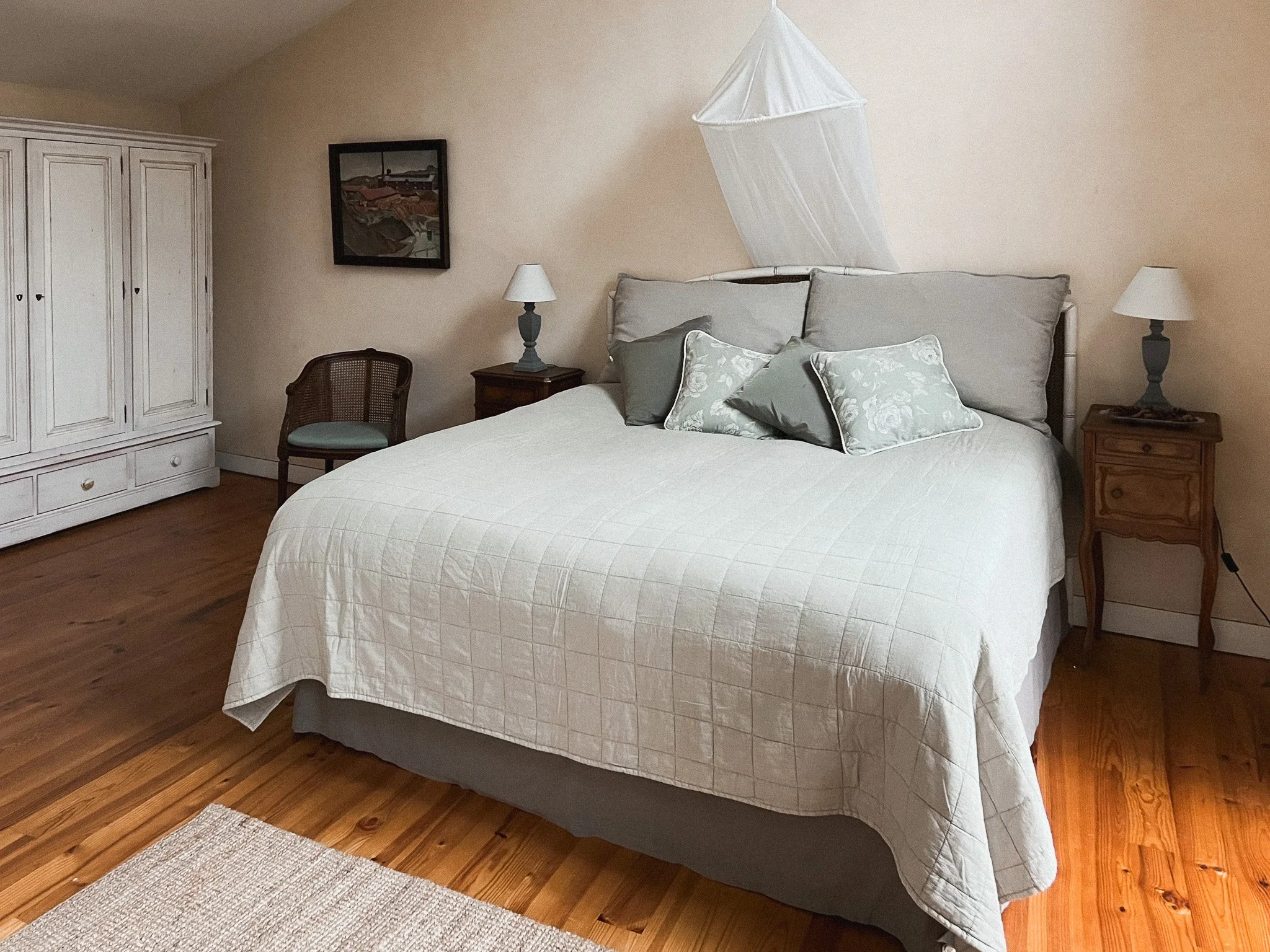 A neatly made bed with white quilt and multiple gray pillows in a cozy bedroom with wooden floors, a wardrobe, a chair, and two nightstands with lamps.