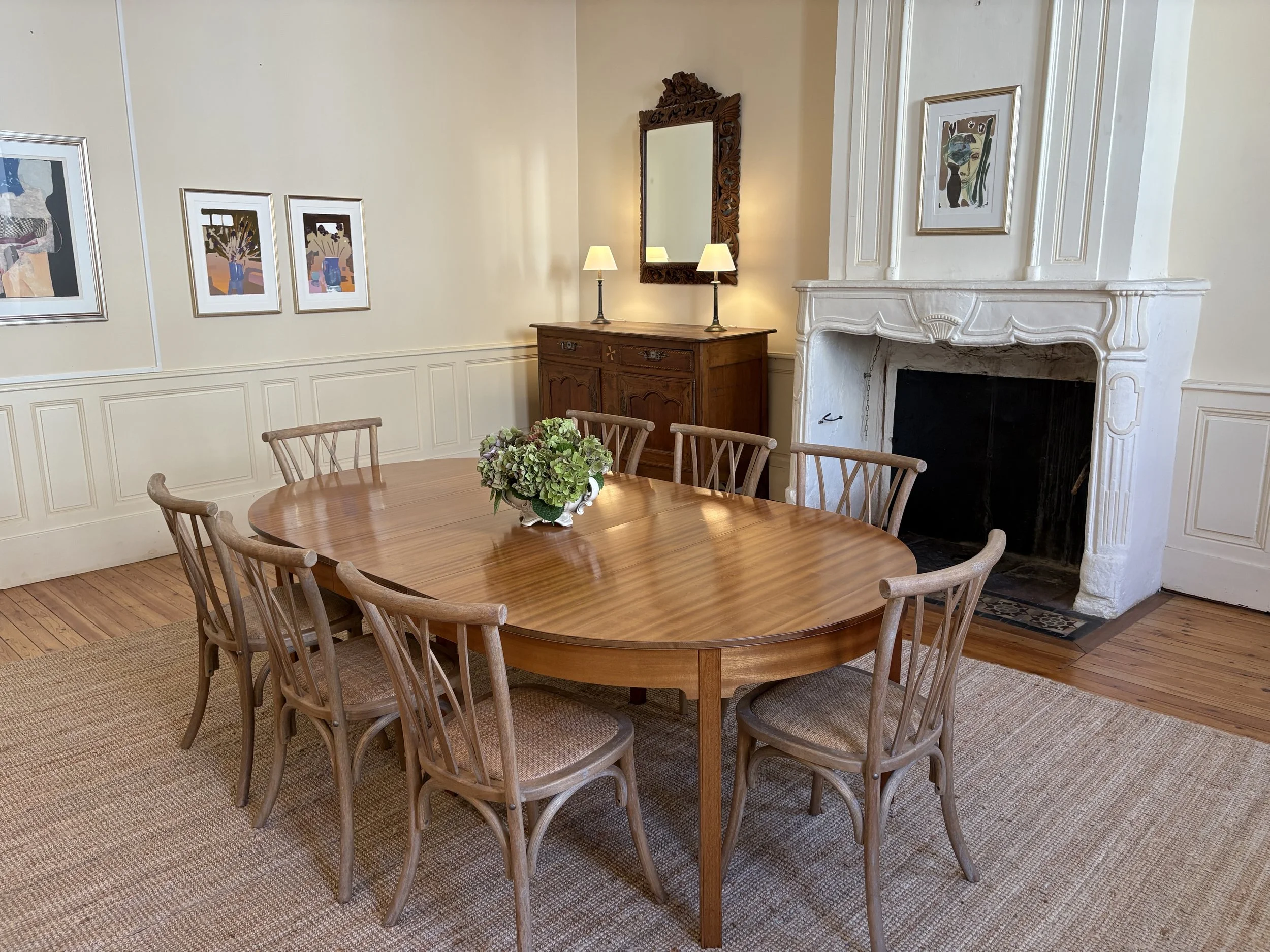 A dining room with a wooden oval table surrounded by eight wooden chairs, a floral centerpiece, a white fireplace, framed artwork on cream-colored walls, a wooden sideboard with two lamps, and a mirror above the sideboard.