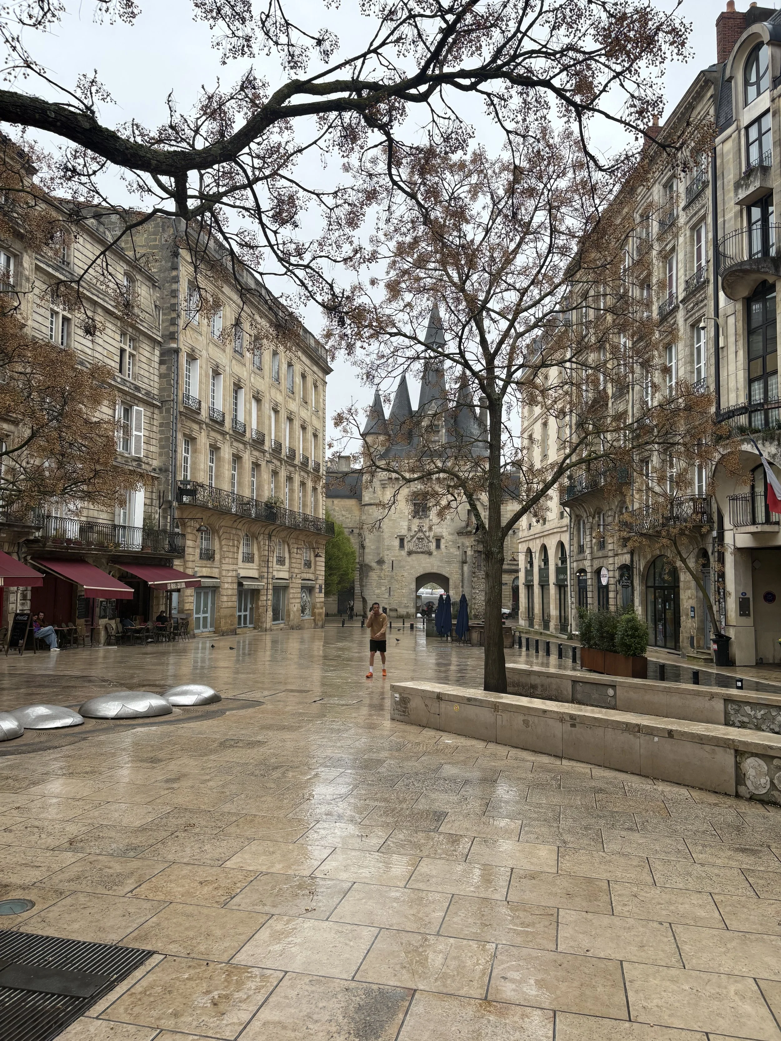 Empty city street with wet pavement, trees, and historic European-style buildings, featuring a stone archway with a castle-like tower in the background.