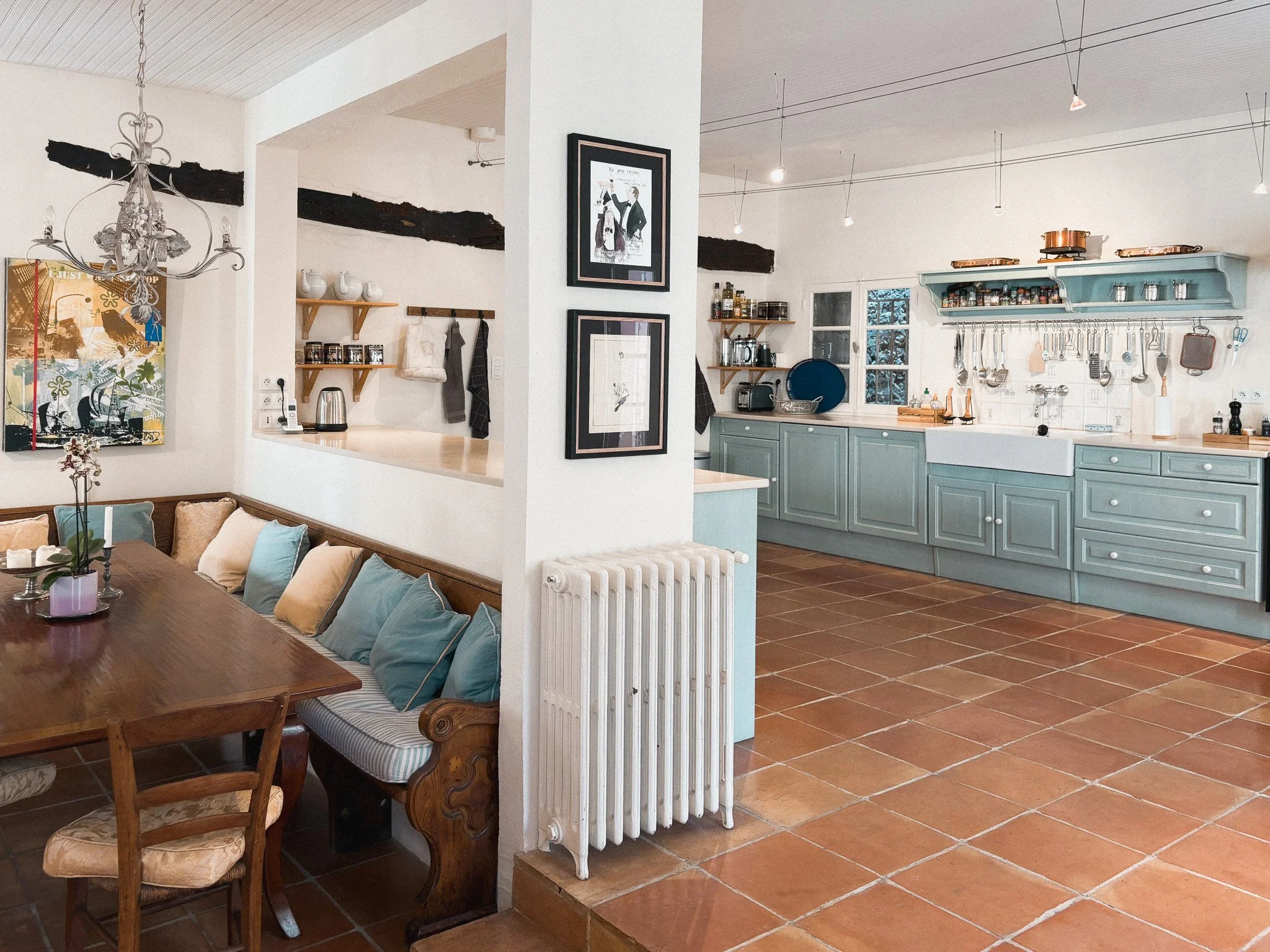 A cozy kitchen and dining area with terracotta tile flooring, light blue cabinets, a white farmhouse sink, and open shelving. Adjacent to it is a dining nook with a wooden table, bench, and chairs, decorated with pillows and a flower arrangement. The