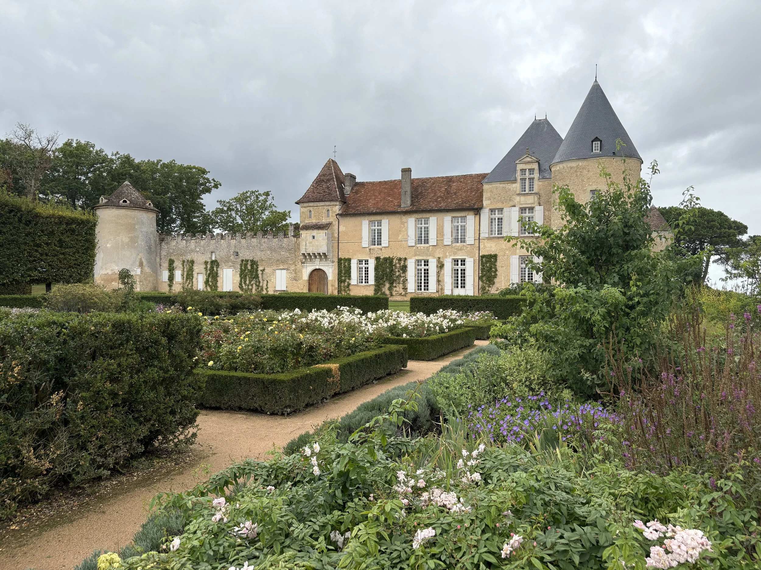 A historic castle with towers and a stone wall, surrounded by a well-maintained garden with colorful flowers and trimmed bushes under an overcast sky.