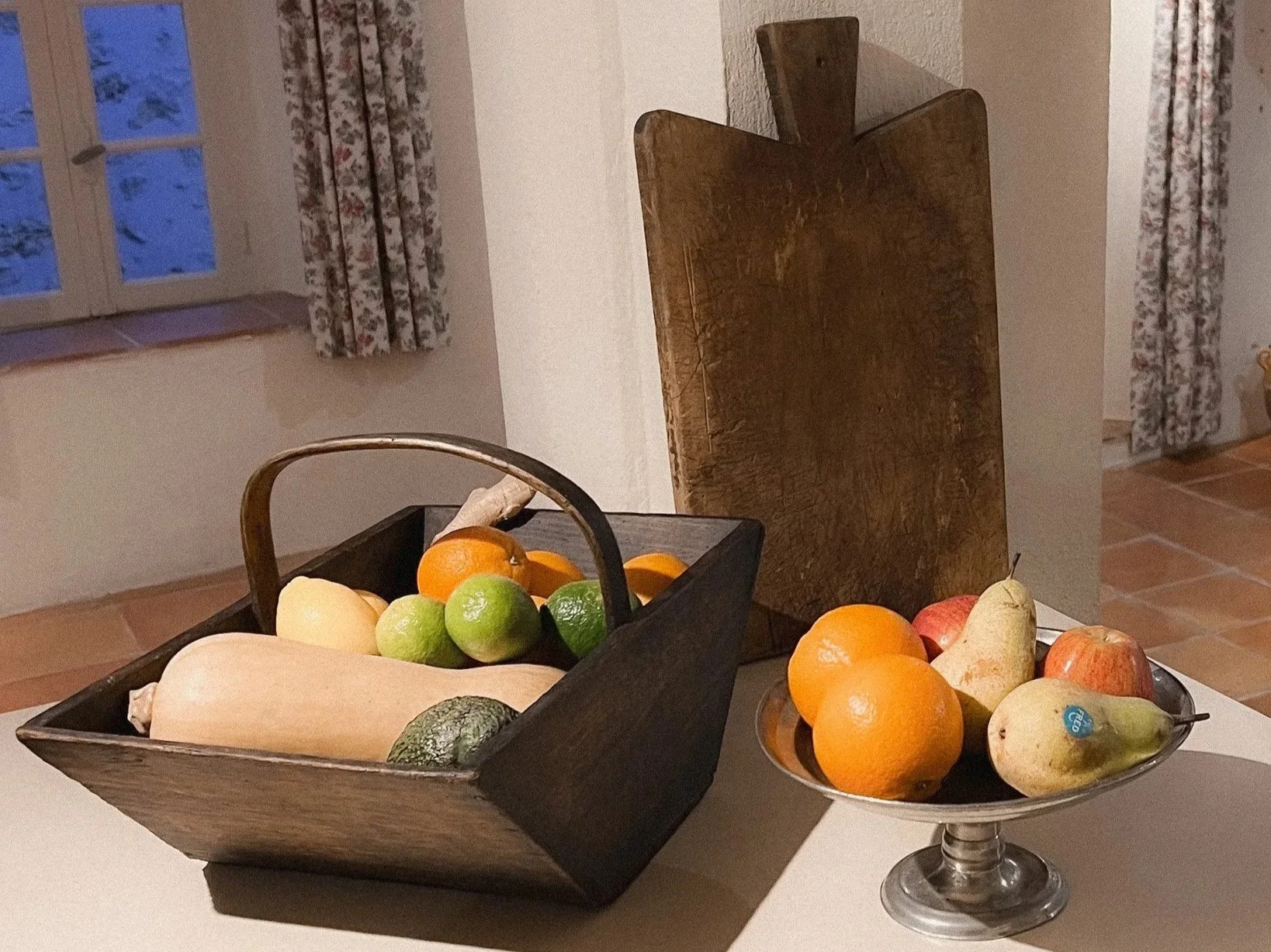 A wooden basket filled with various fruits including apples, oranges, lemons, a pear, and a squash. Next to it, a silver pedestal bowl holds additional fruits such as oranges, a pear, and an apple. Behind the basket, a wooden cutting board leans against the wall, with a window with floral curtains visible in the background.
