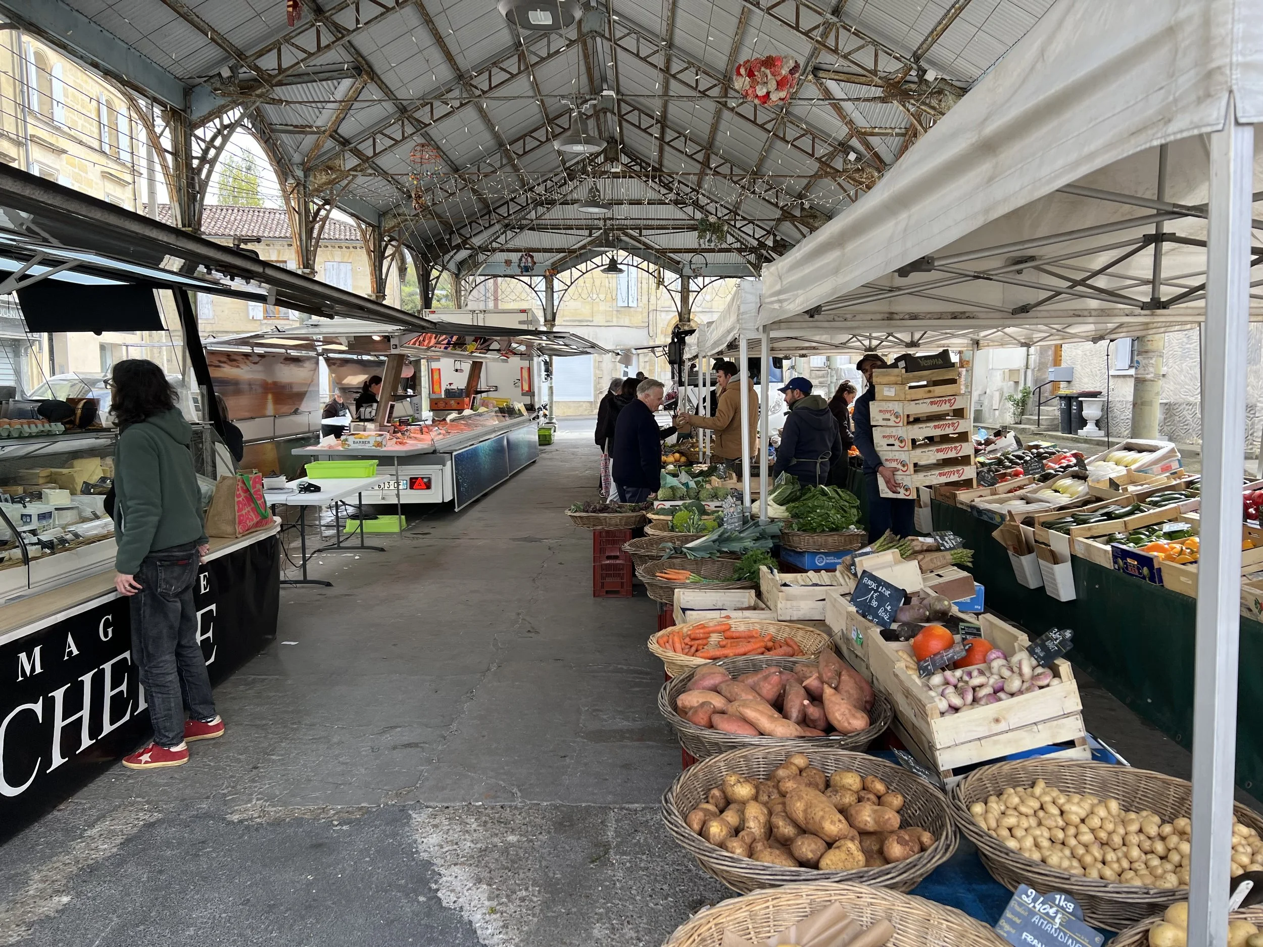 An indoor farmers market with stalls selling fresh vegetables and seafood, beneath a metal arched ceiling with hanging decorations.