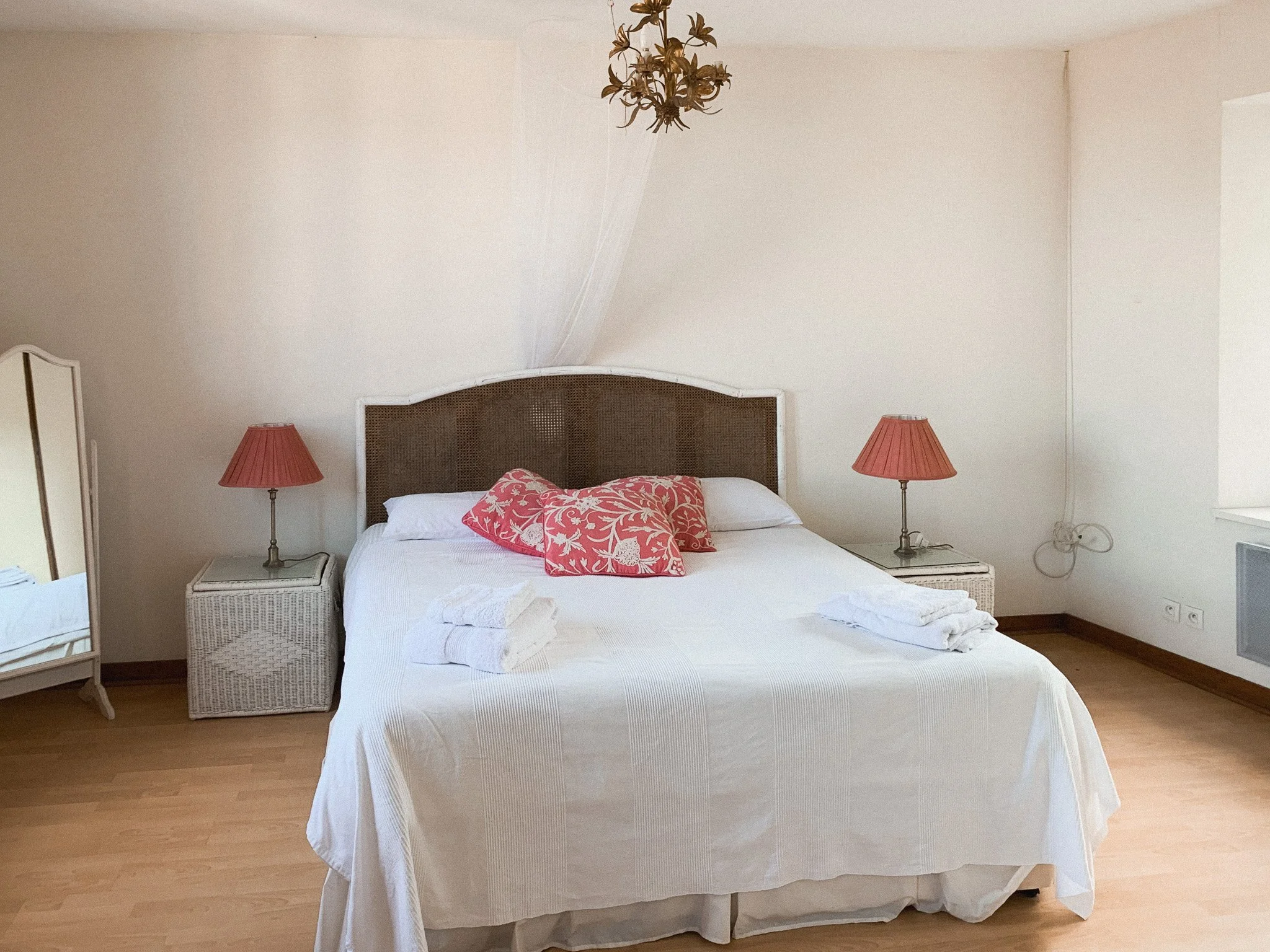 A bedroom with a large bed covered in white bedding, two stacked pink pillows, and two folded towels on the bed. There are two small nightstands with pink lamps on each side. A mirror and chandelier are also visible in the room.