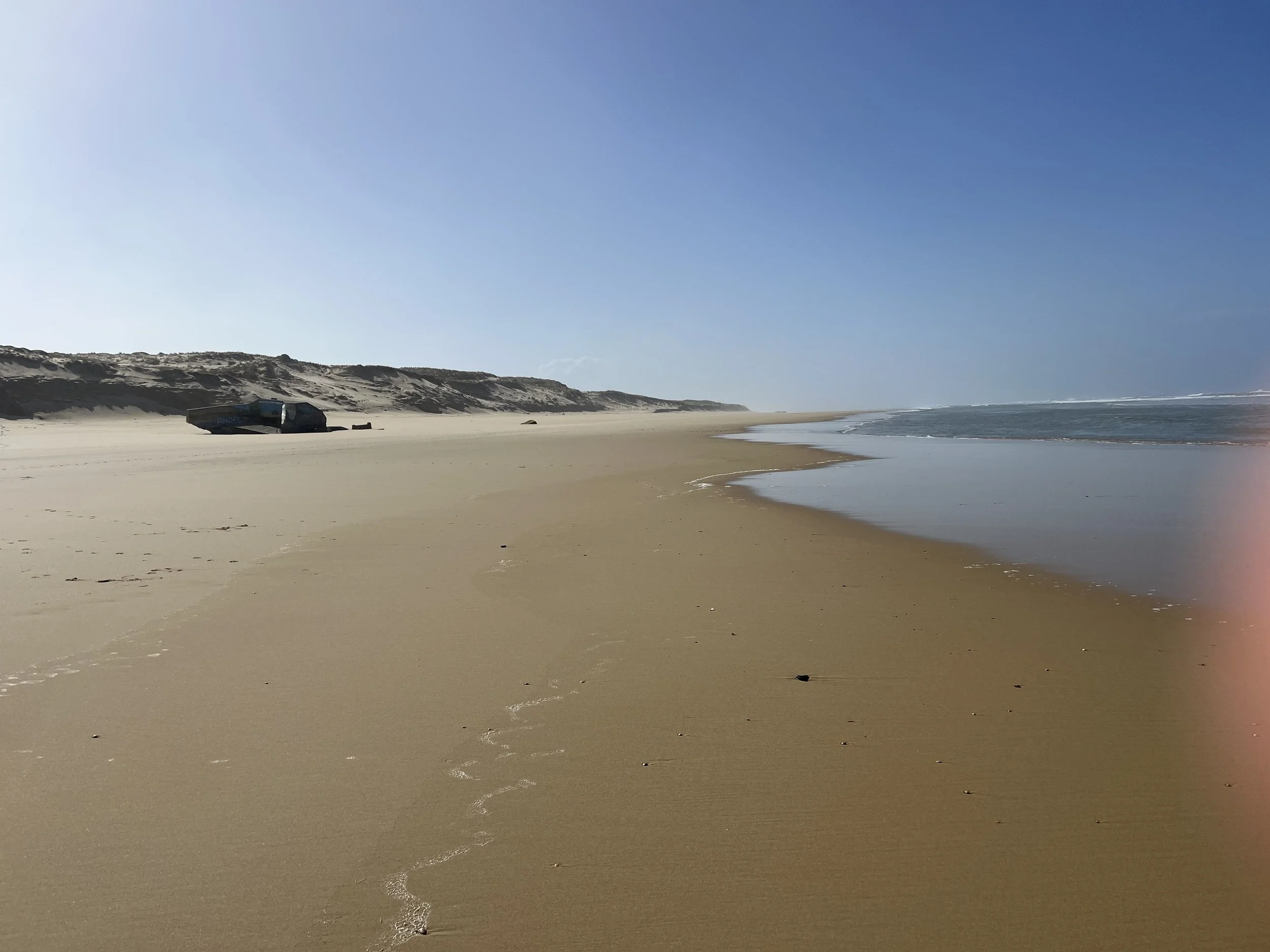 Empty sandy beach with some trash and a deteriorated bus on the sand, dunes in the background, cloudy sky, and the ocean on the right.
