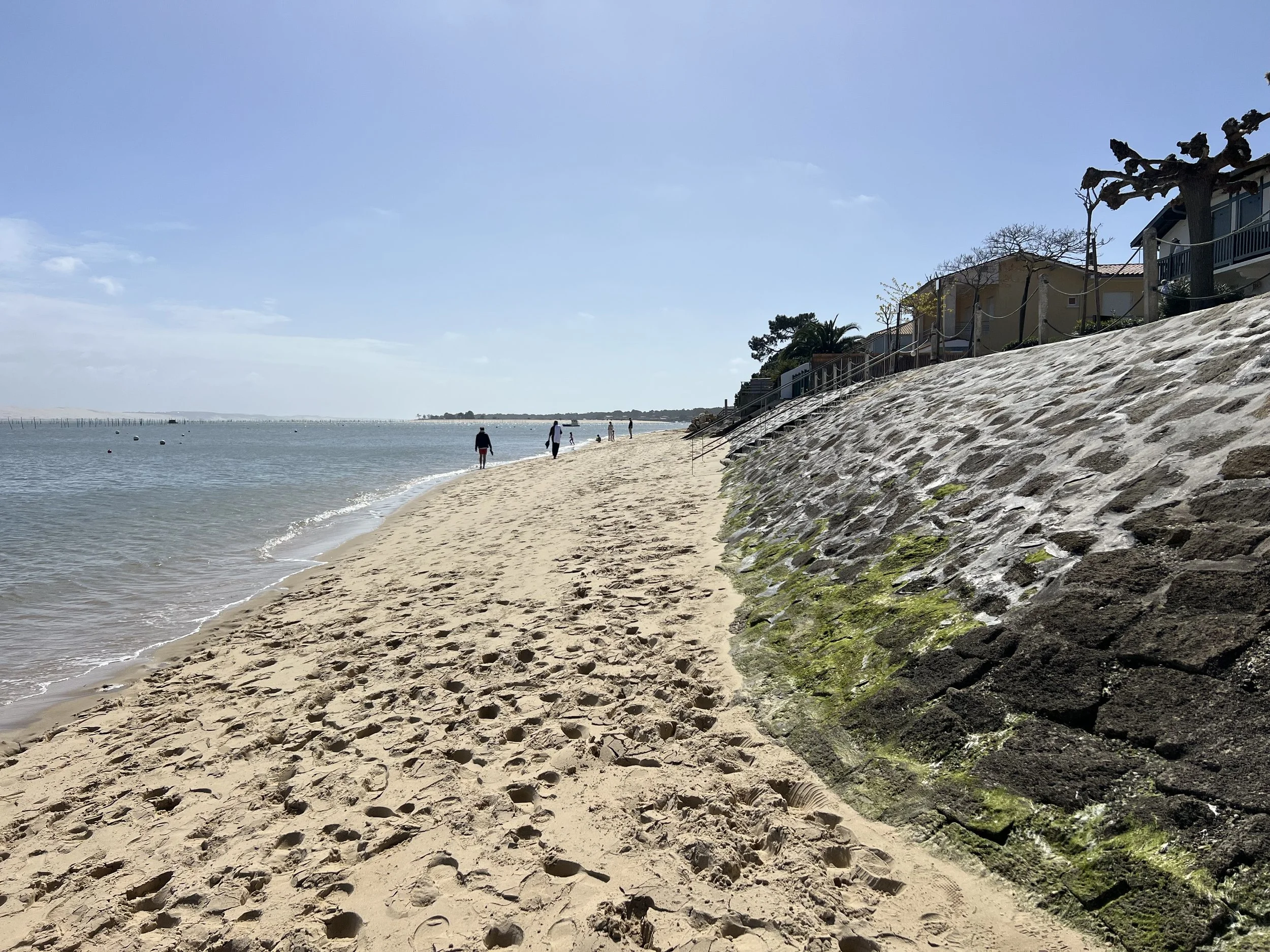 A sandy beach with footprints, calm water on the left, and houses on a stone embankment on the right, under a clear blue sky.