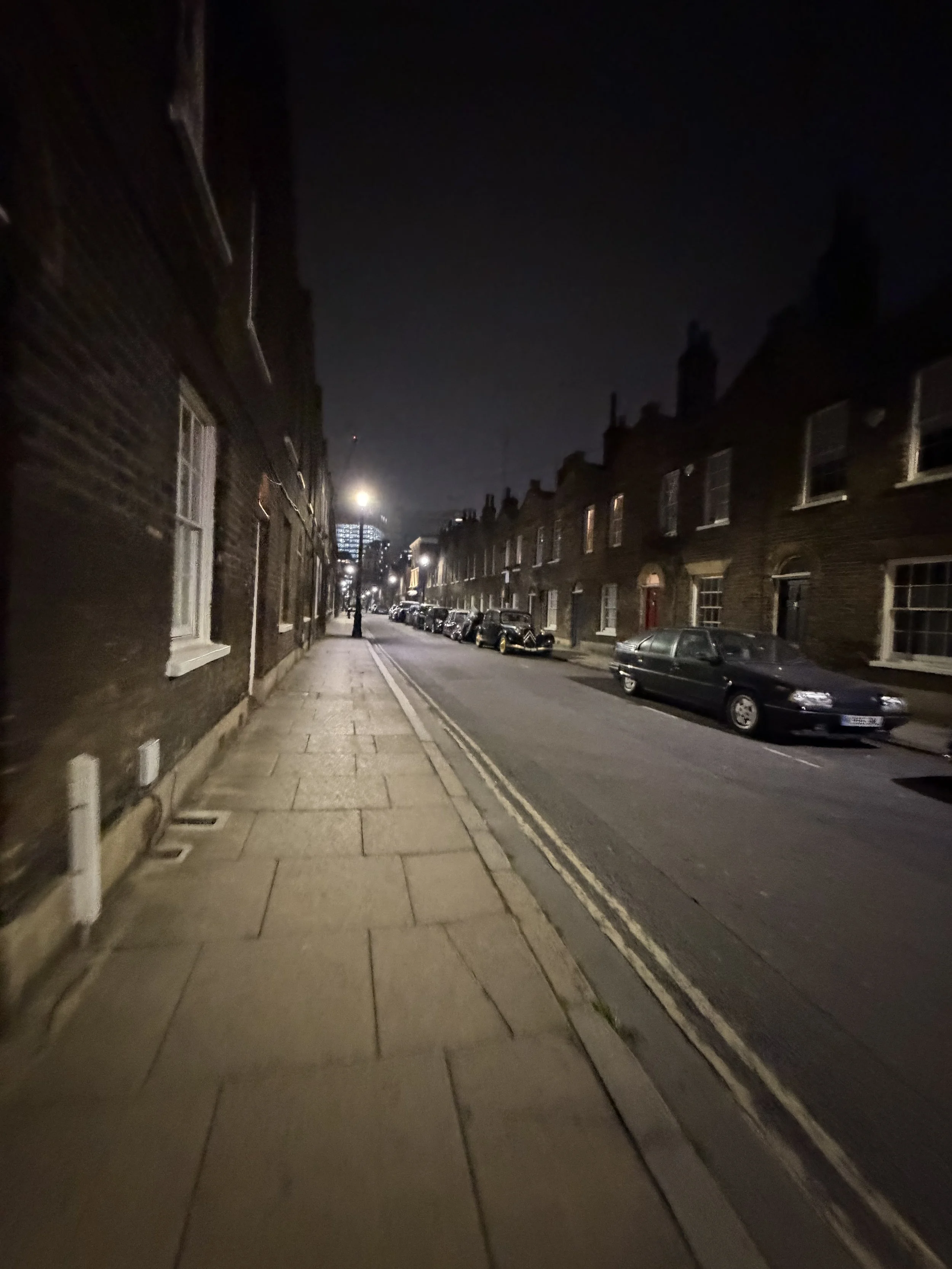 A nighttime street scene with a sidewalk on the left and parked cars along the right side of the street. There are old brick buildings on both sides, and the street is illuminated by a few streetlights.