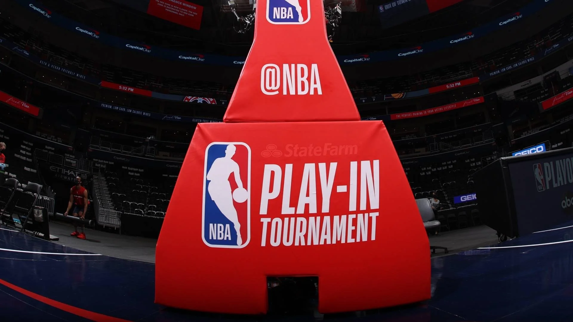 Basketball court with a large red pole in the foreground displaying the NBA Play-In Tournament logo and social media handle @NBA, with empty seats and a few athletes in the background.
