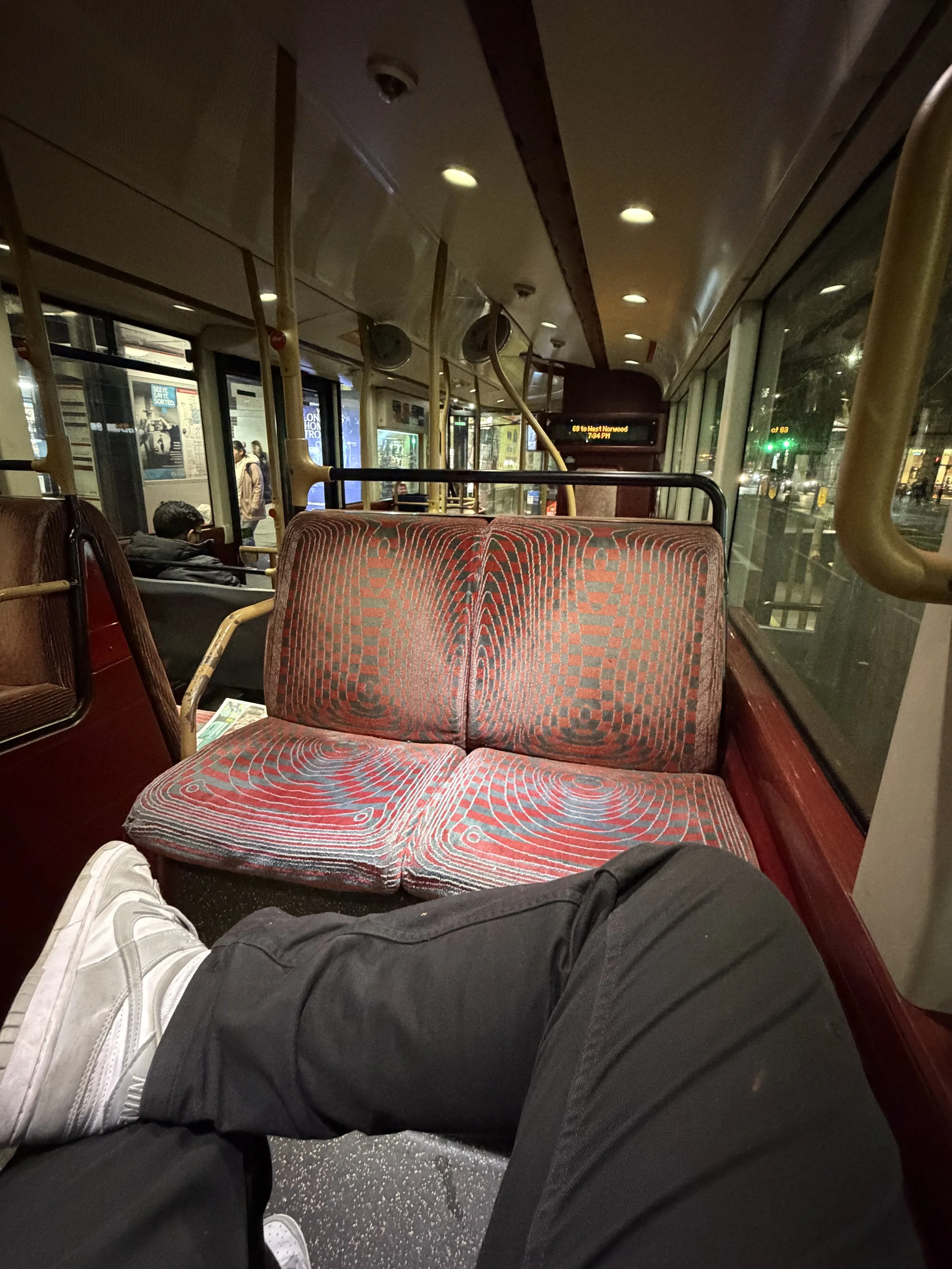 View inside a vintage bus showing red patterned seats, some passengers, and a city street through large windows at night.