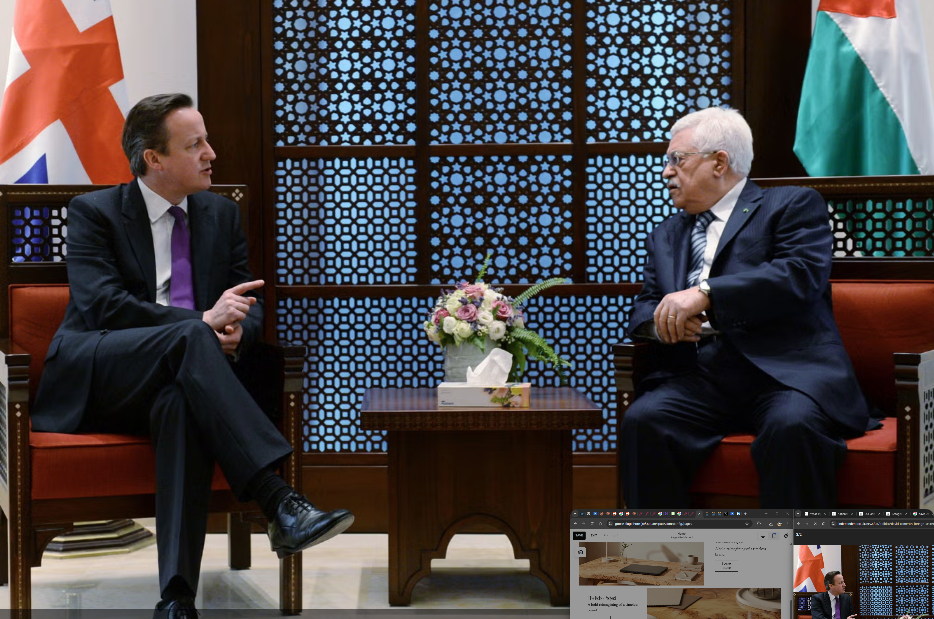 Two men in suits having a conversation in a formal setting with flags and a decorative screen in the background. One man has dark hair and is wearing a purple tie, while the other has white hair and is wearing a patterned tie. A small table with a bouquet of flowers and a box of tissues is between them.