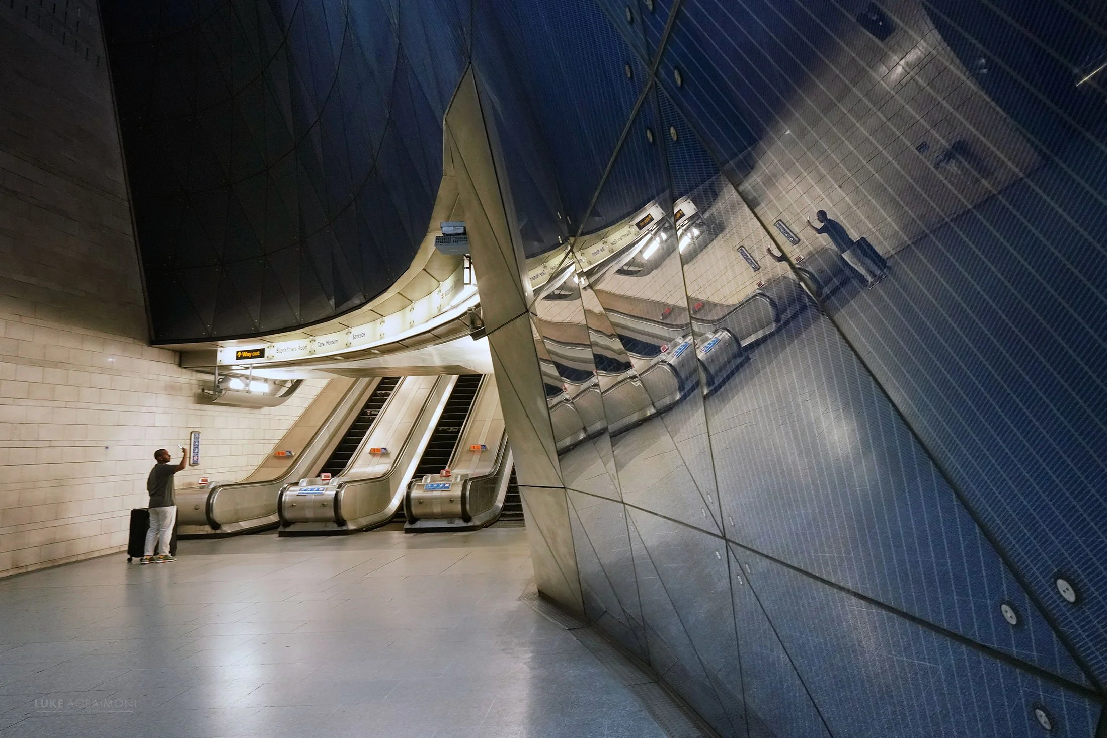 An underground subway station with escalators and a reflective wall. A person is standing near the escalators, taking a photo or looking at their phone.
