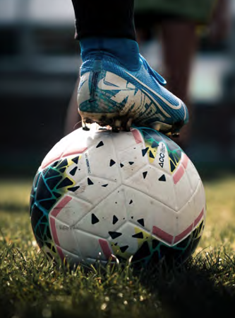 Close-up of a person wearing blue soccer shoes balancing a white soccer ball with colorful geometric patterns on grass.