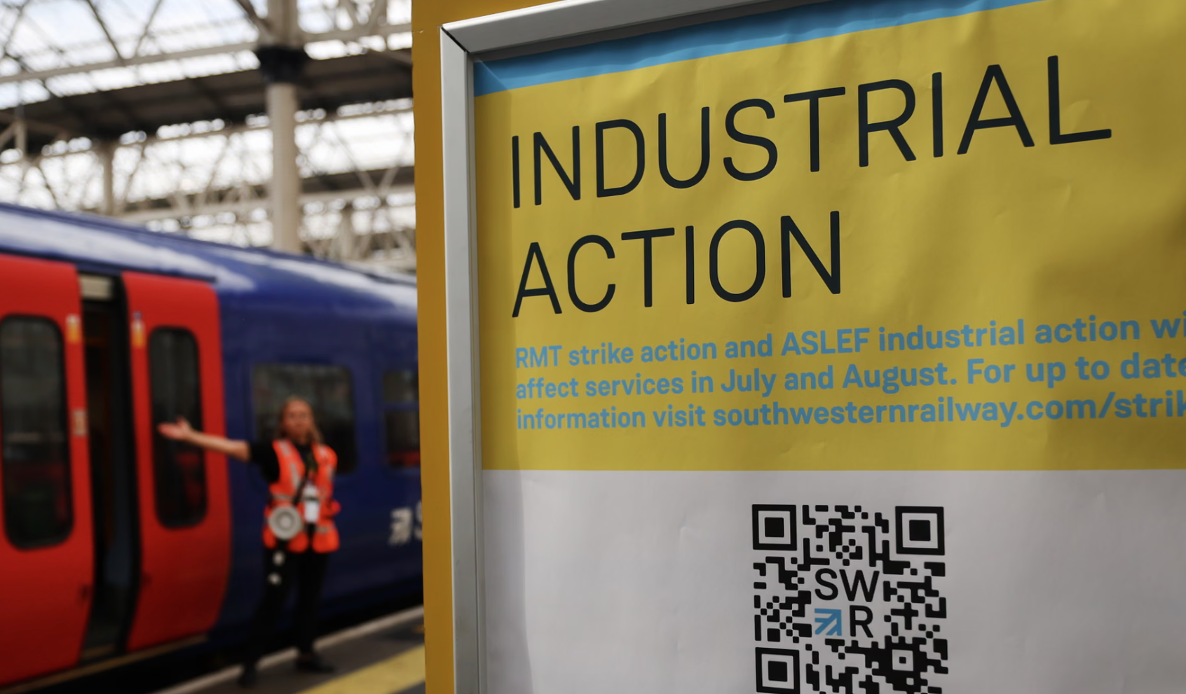 Sign indicating industrial action affecting train services, with a person in a safety vest in the background near a blue and red train.