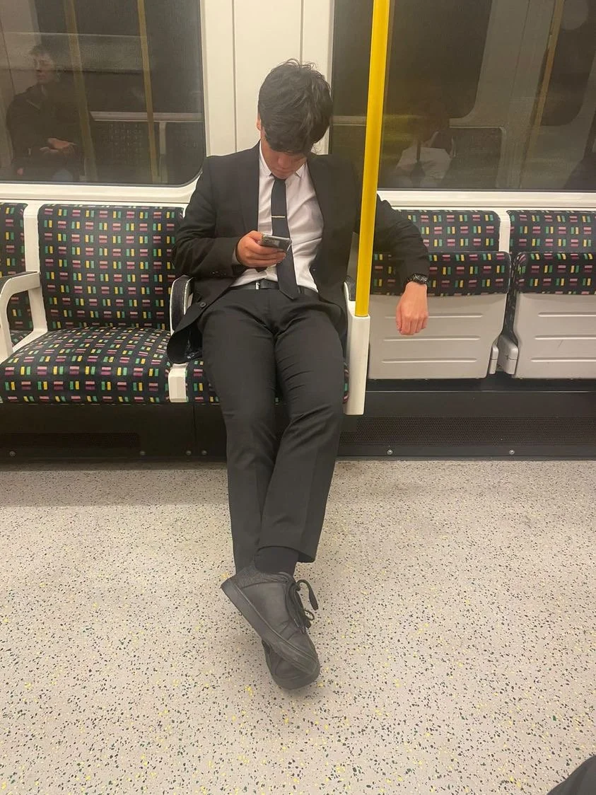A young man dressed in a black suit and tie sitting on a train, looking at his phone with one foot resting on the floor.