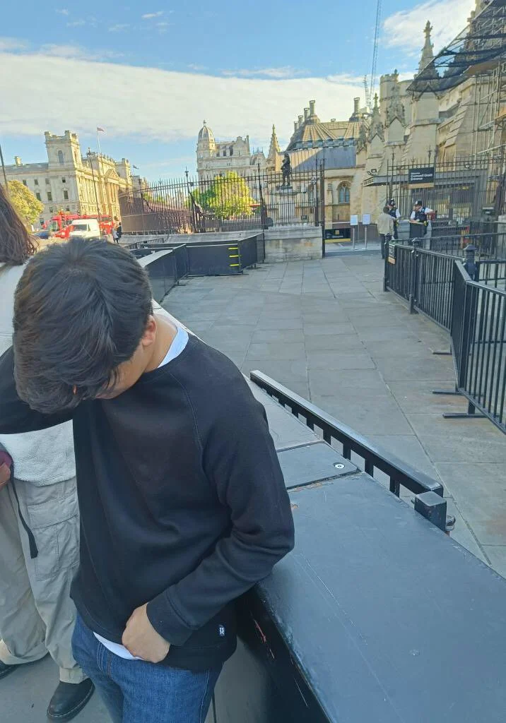 Young man wearing a black jacket and blue jeans, looking down, standing outdoors near a historic building.