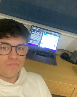 A young man wearing glasses and a white hoodie taking a selfie in front of a laptop on a wooden desk. The laptop screen displays a coding interface and a messaging app.
