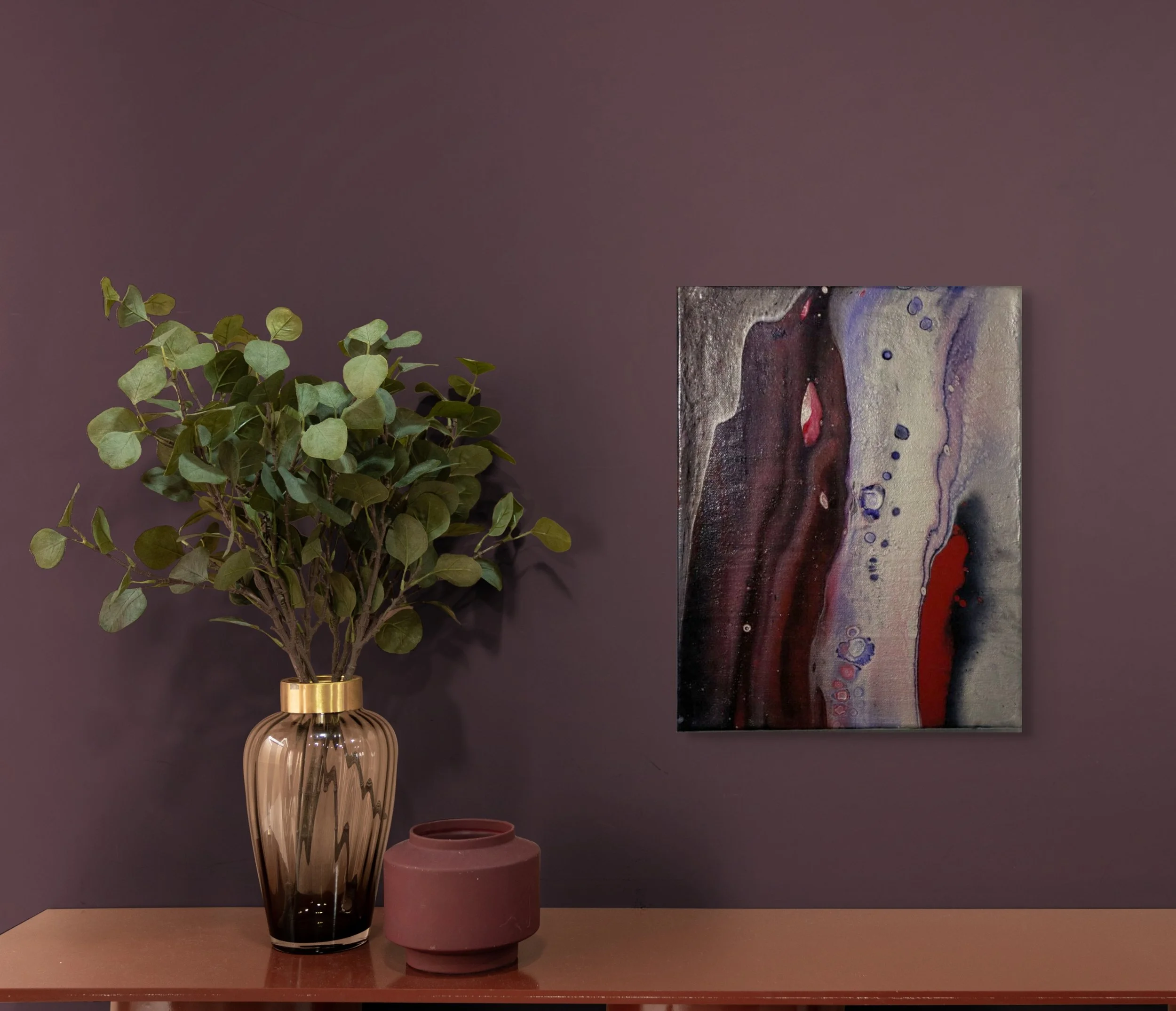 A decorative arrangement featuring a tall glass vase with green leafy plant, an abstract painting on mauve wall, and two small pink vases on a wooden surface.