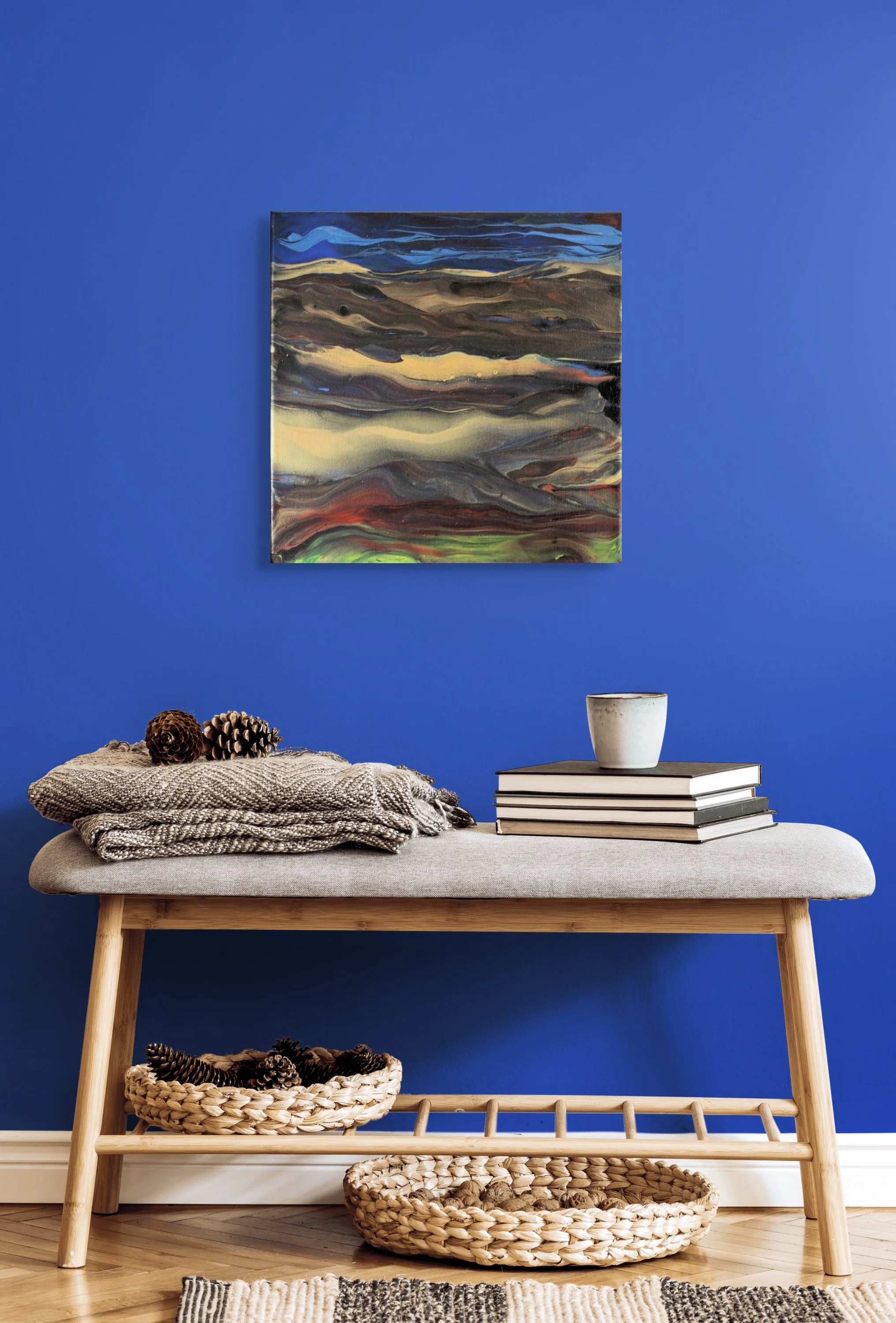 A wooden furniture piece with three shelves, decorated with baskets of pinecones, a stack of books with a white cup on top, and decorated with a throw blanket and a small rug, set against a vibrant blue wall with a colorful abstract painting.
