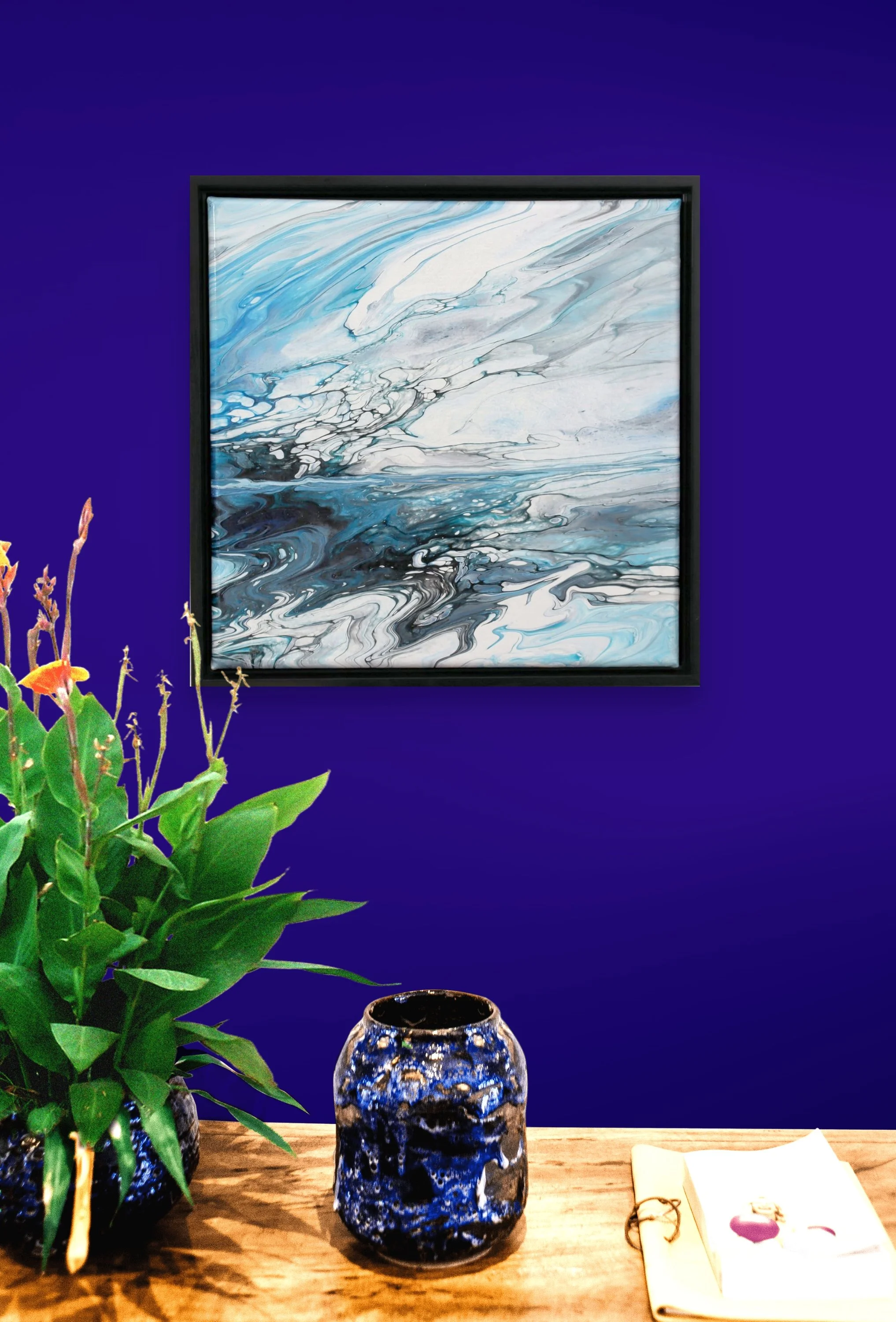 Decorative interior scene with a blue and black marbled vase, green potted plant, framed abstract art with blue and white swirling patterns on a deep blue wall, and a ceramic vase on a wooden surface with a notebook and gold ring nearby.