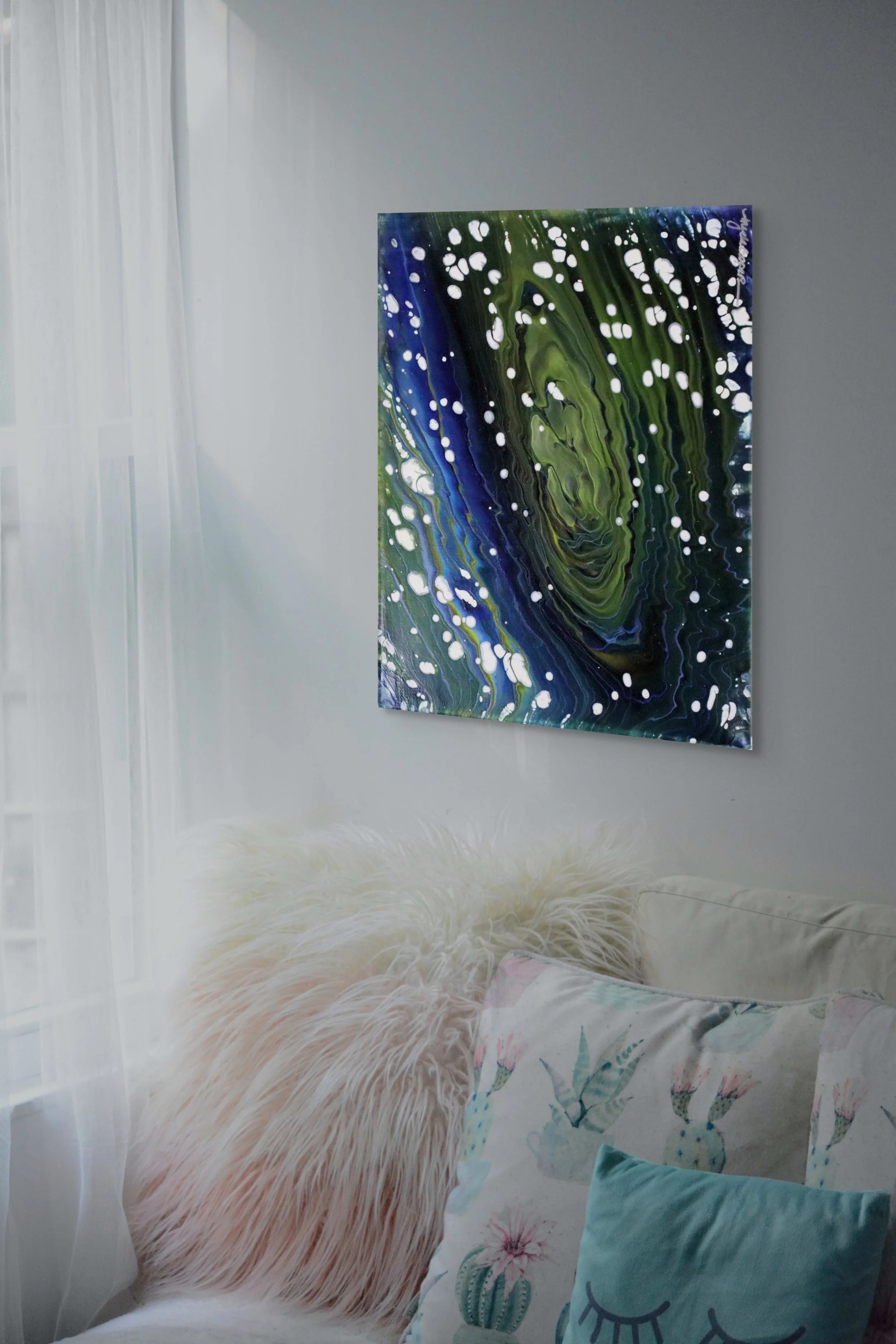 A colorful abstract painting hanging on a white wall in a room with curtains. In front, there is a fluffy beige pillow, a sheet with floral and cactus prints, and a light blue pillow with a smiling face.